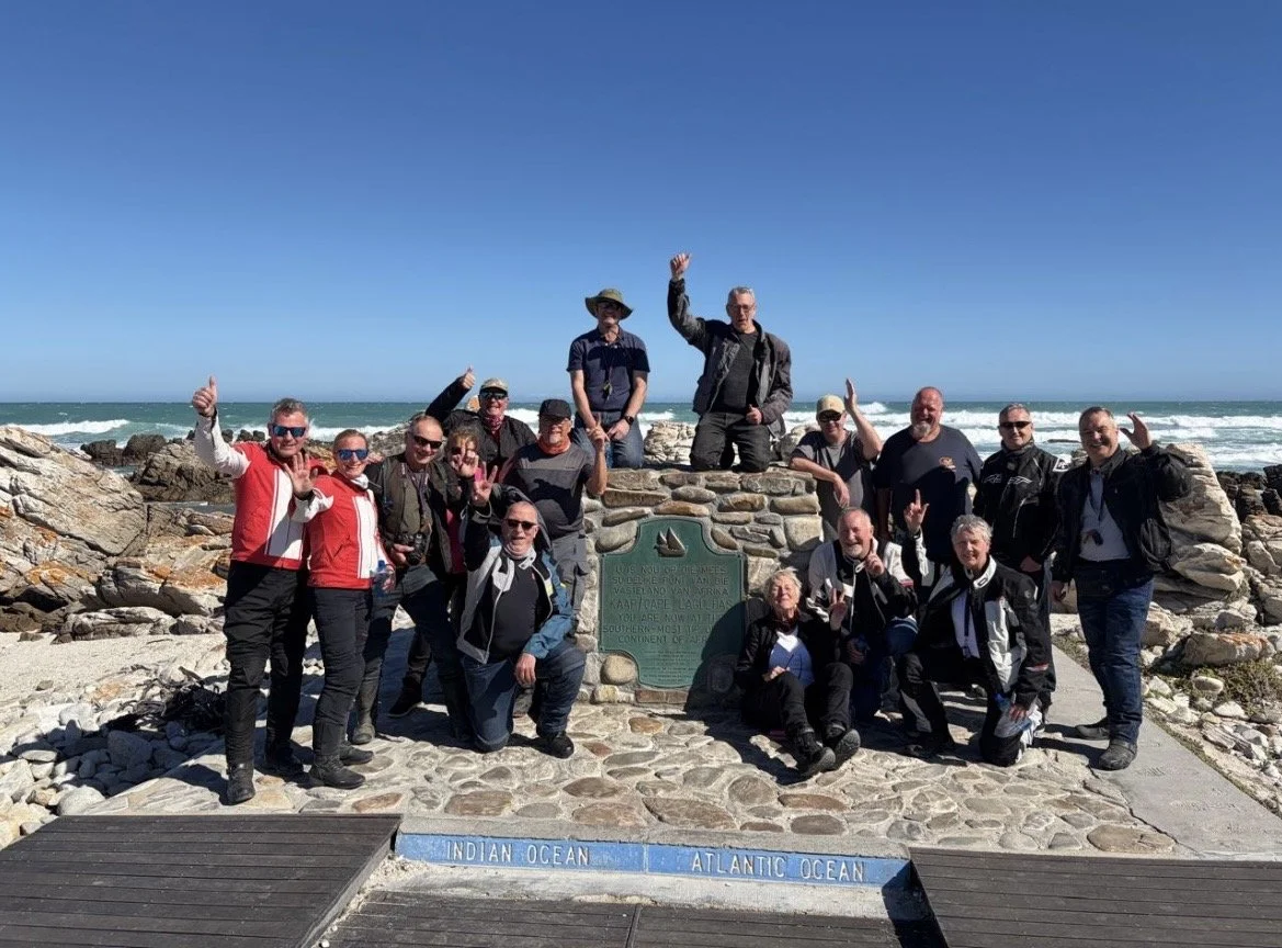 Group of people posing at the confluence of the Indian Ocean and Atlantic Ocean, with a blue sky and ocean waves in the background.