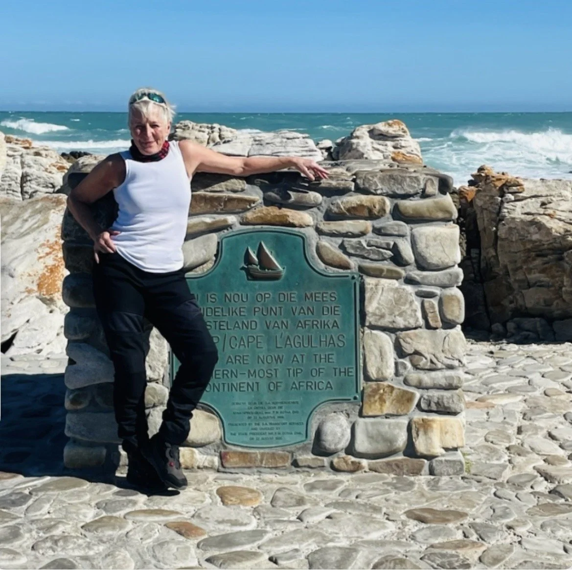 Older woman standing near a stone marker by the ocean at Cape Agulhas, South Africa, with waves in the background.