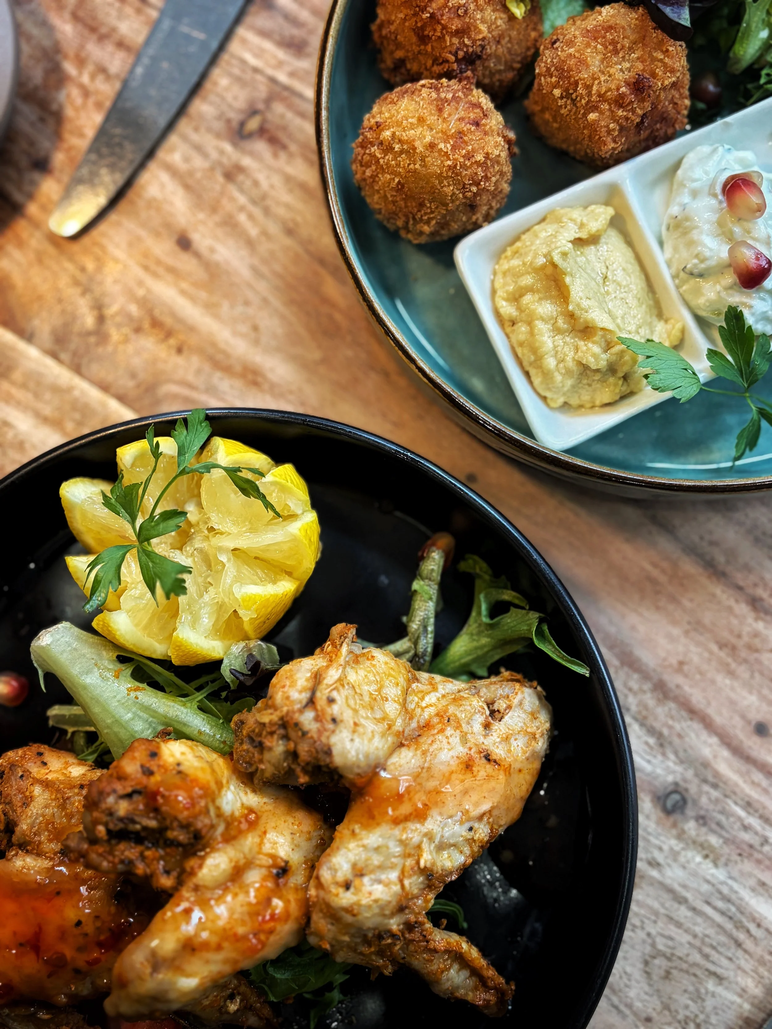 Close-up of a bowl with fried chicken and prawn, garnished with greens, with a lemon wedge on a separate dish, and croquettes with dips in the background.