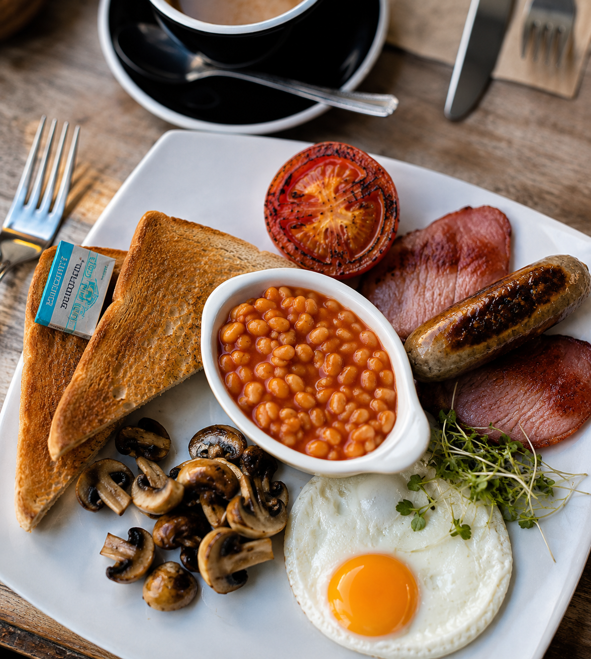 Full English breakfast with toasted bread, baked beans, grilled tomato, sautéed mushrooms, fried egg, bacon, sausage, and microgreens on a white plate, with a cup of black coffee in the background.