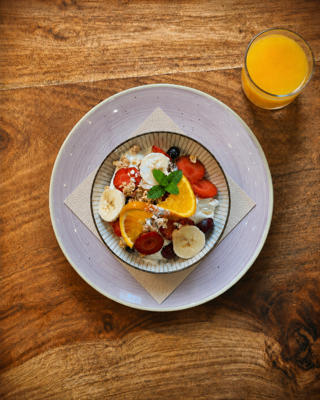 A bowl of yogurt topped with sliced bananas, strawberries, orange slices, blueberries, grapes, chopped nuts, and mint leaves, accompanied by a glass of orange juice on a wooden table.