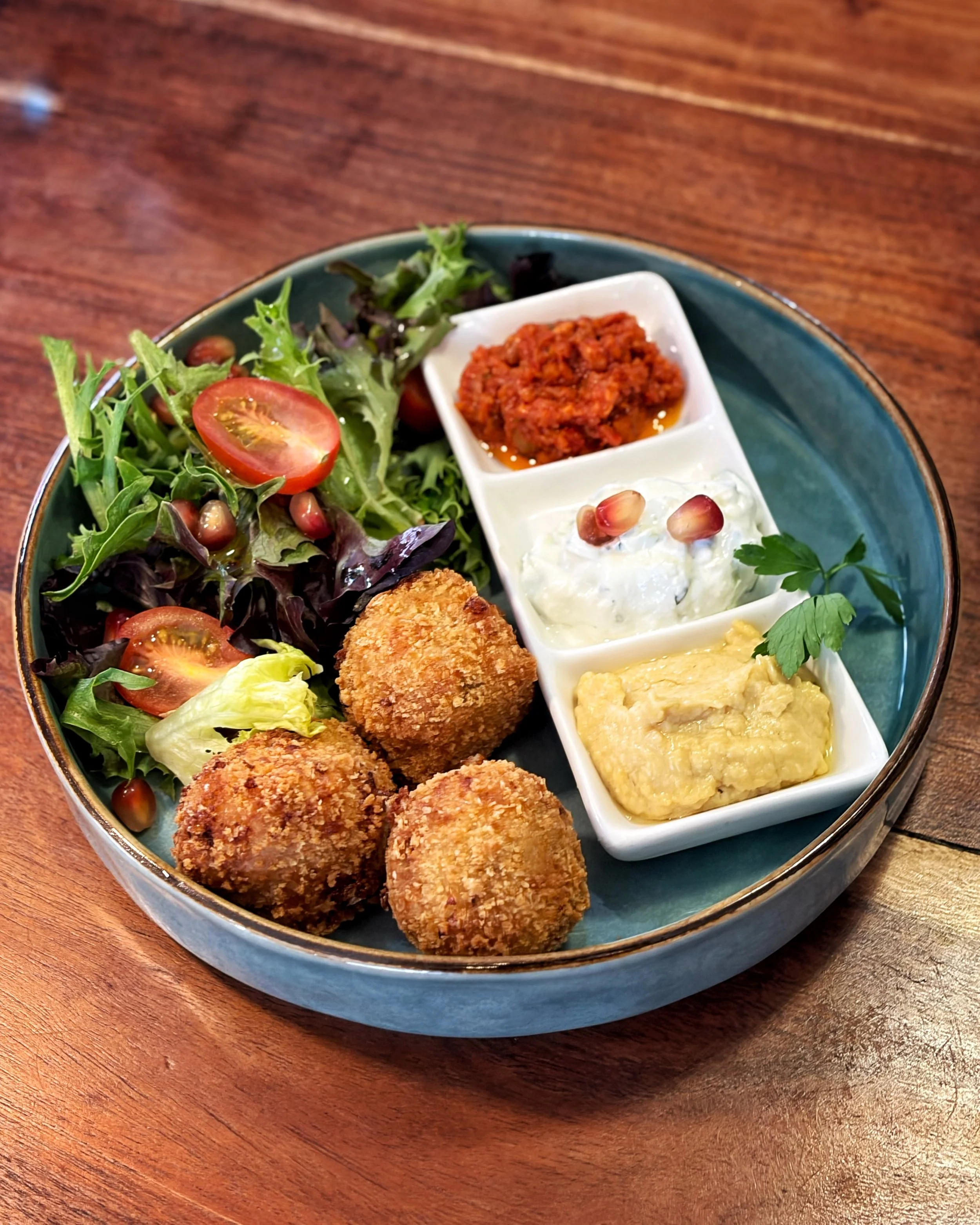 A plate with a mixed salad, three fried croquettes, and three small dishes containing red sauce, sour cream or yogurt with pomegranate seeds, and yellowish egg salad, set on a wooden table.