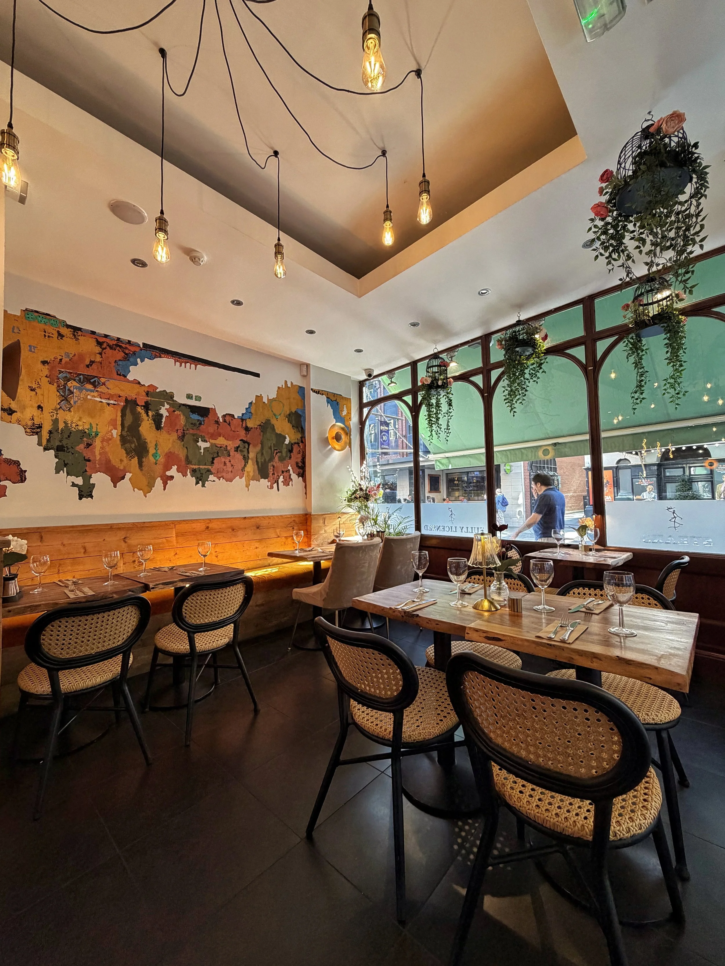 Interior of a cozy restaurant with wooden tables, woven chairs, hanging light bulbs, a colorful abstract mural on the wall, large windows with hanging plants, and a street scene outside.