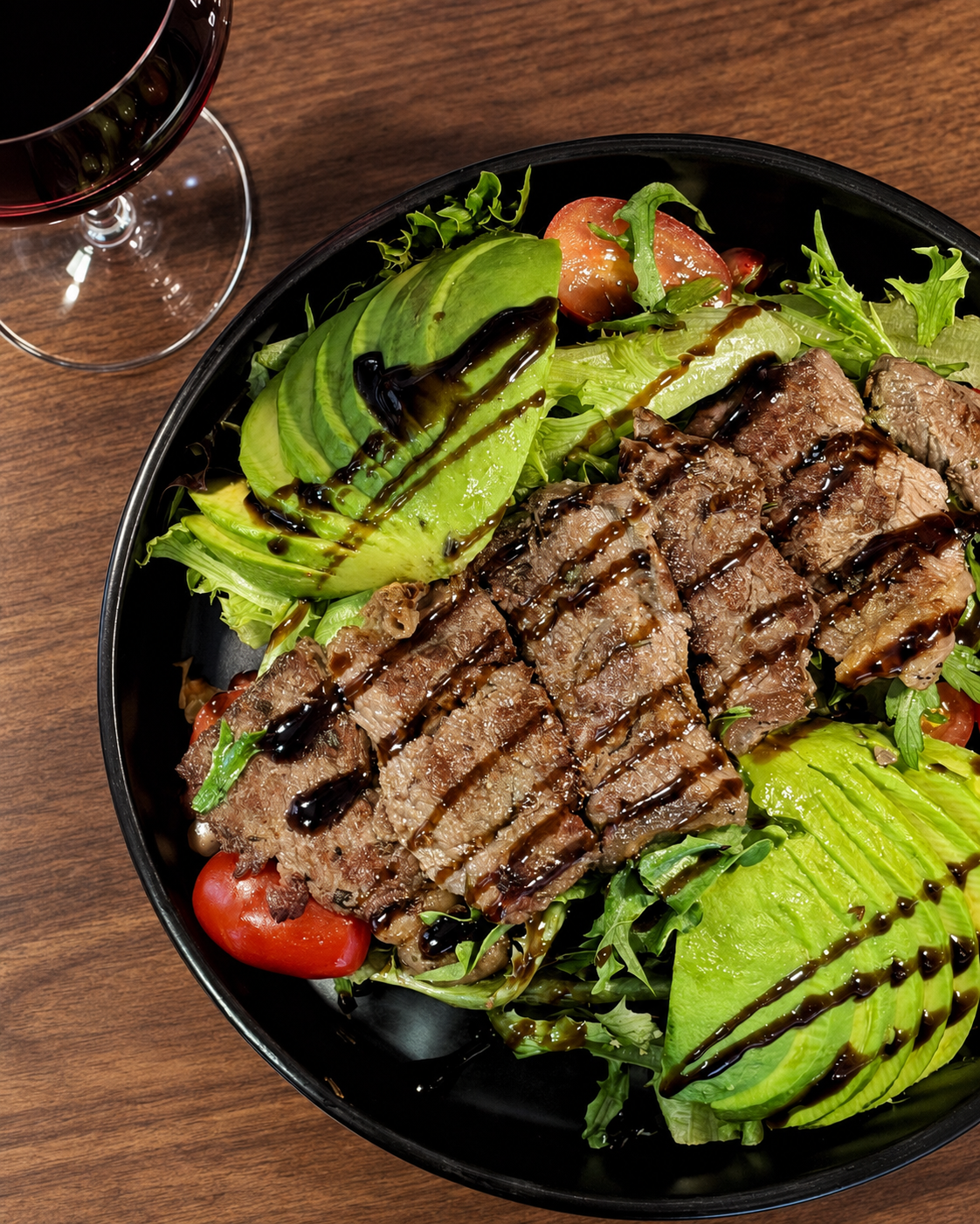 Salad with sliced avocado, cherry tomatoes, grilled steak slices, mixed greens, drizzled with balsamic glaze, and a glass of red wine