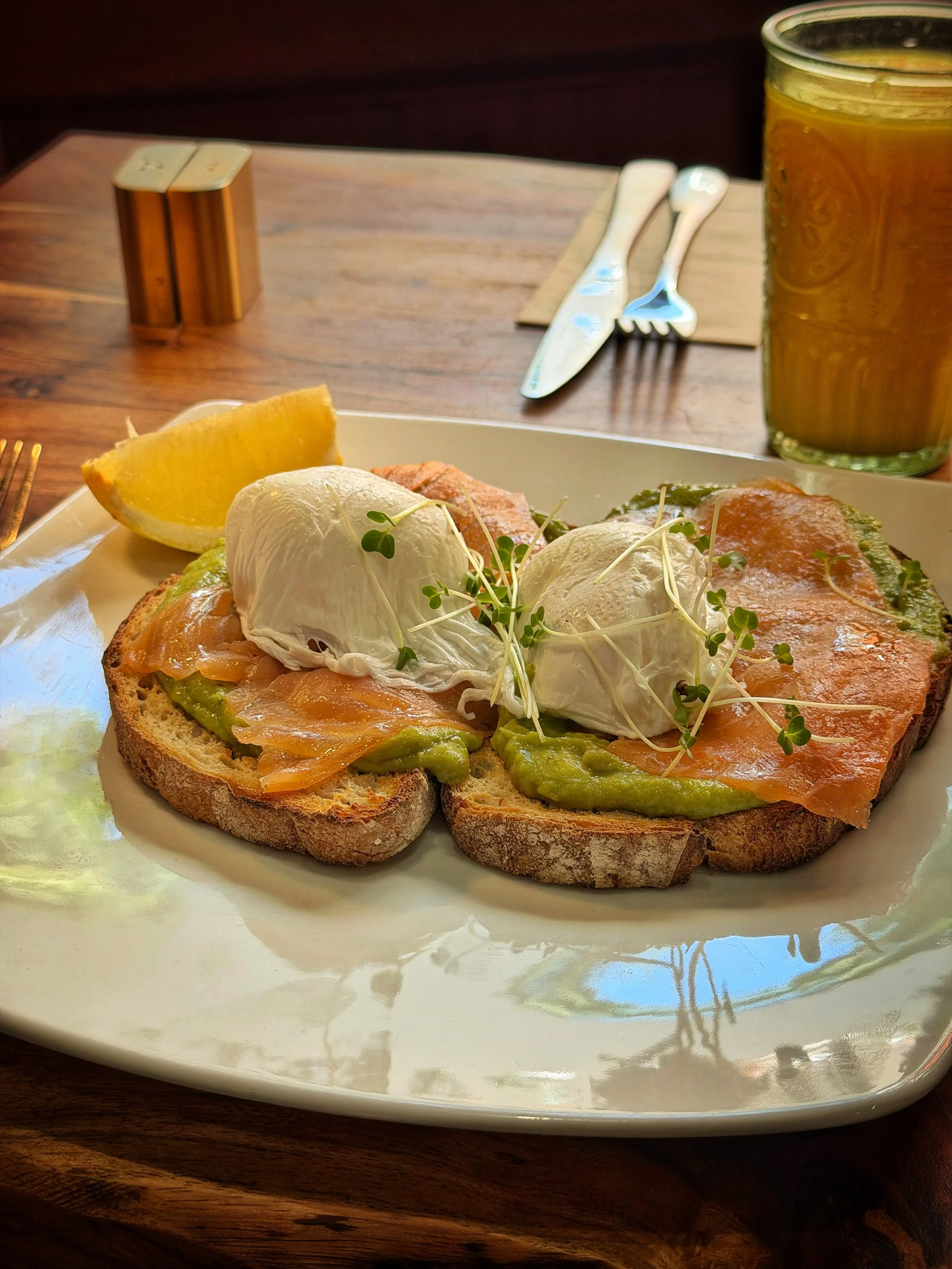 Open-faced toast with smoked salmon, poached eggs, avocado spread, and microgreens on a white plate, with lemon wedge, salt and pepper shakers, and cutlery on a wooden table.