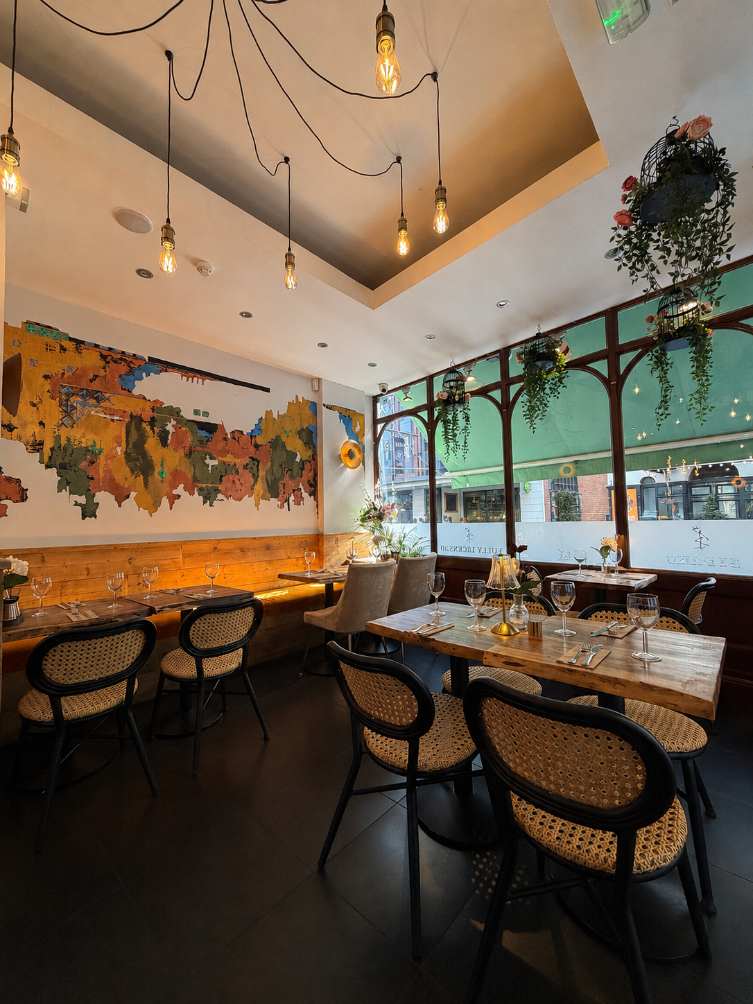 Inside a cozy restaurant with wooden tables, upholstered chairs, hanging light bulbs, artwork on the wall, and large windows with hanging plants.
