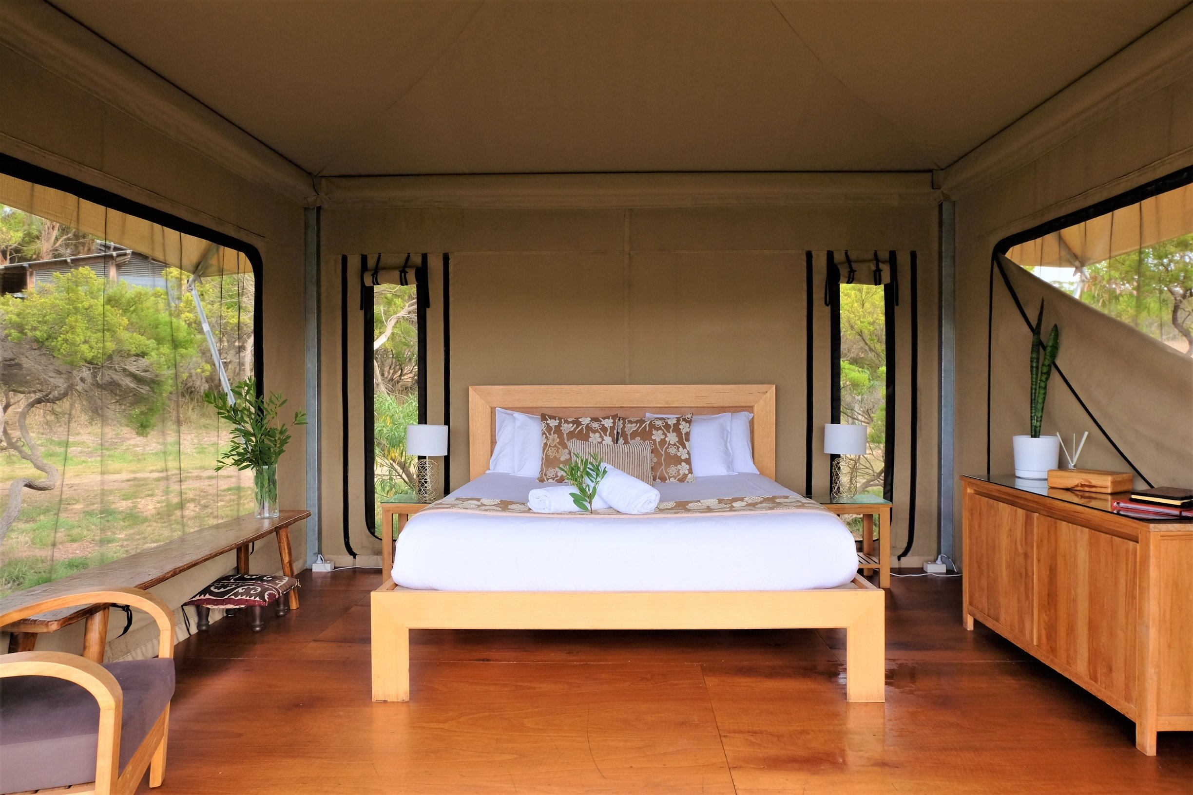 Interior of a cozy tent with a bed, decorative pillows, and side tables with lamps, surrounded by greenery visible through the tent's windows.