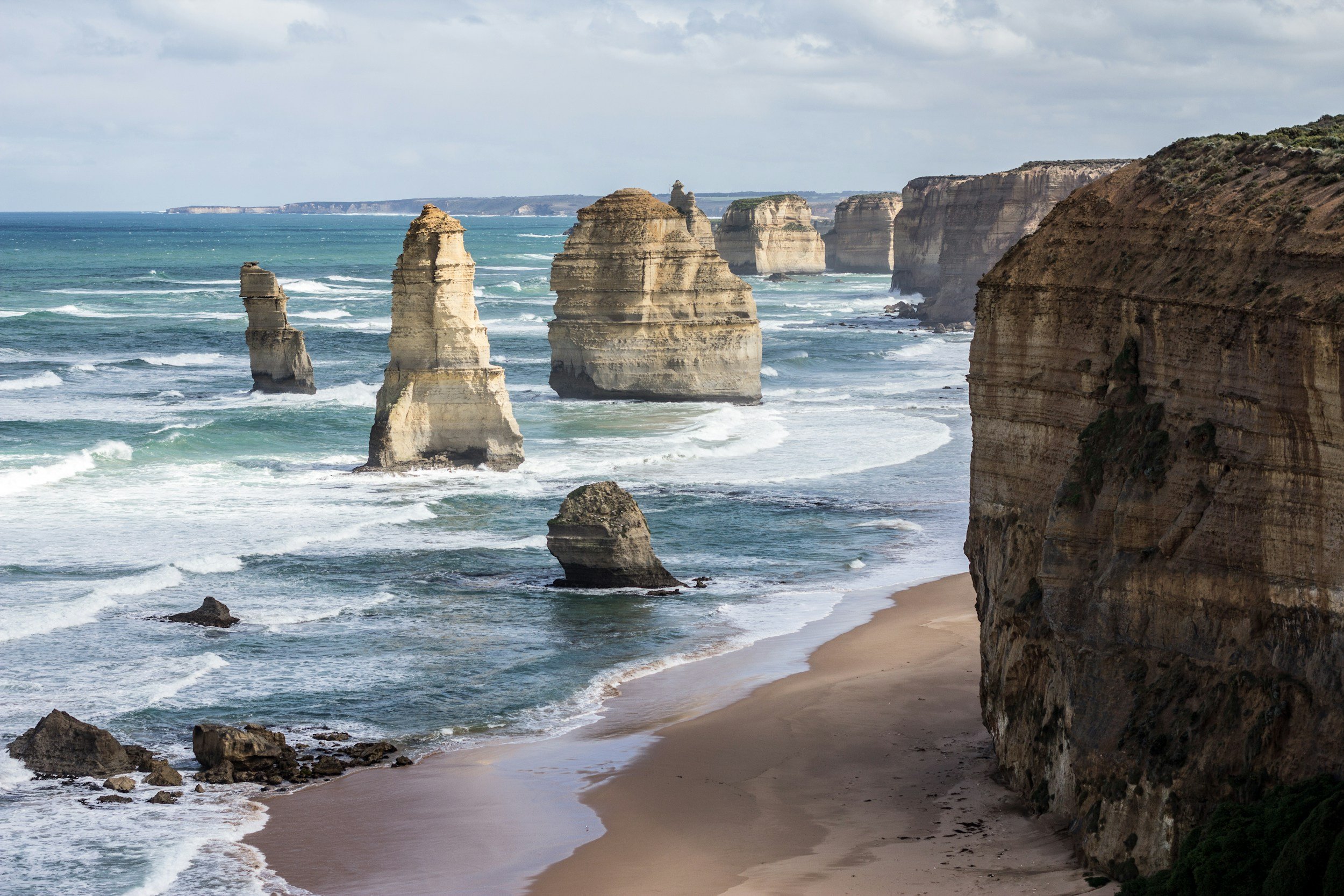 Cliffs and rock formations along a beach with waves crashing, under a partly cloudy sky.