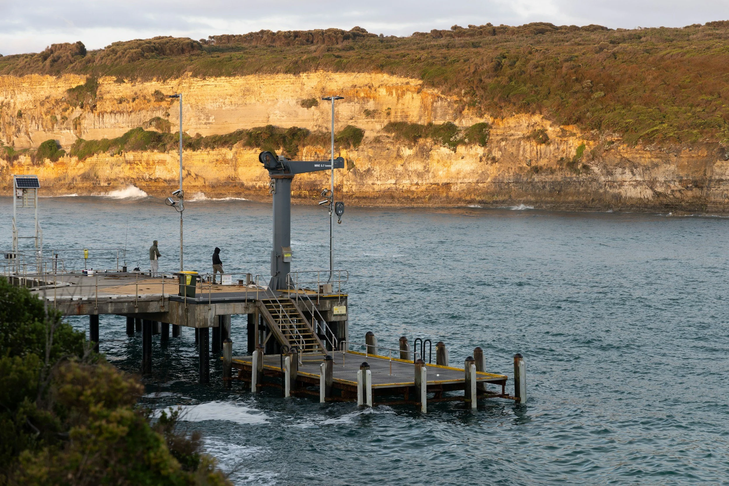 A waterside pier with some people standing on it, equipped with weather instruments. Behind is a large yellow and brown rocky cliffside with vegetation on top, and water with small waves below.