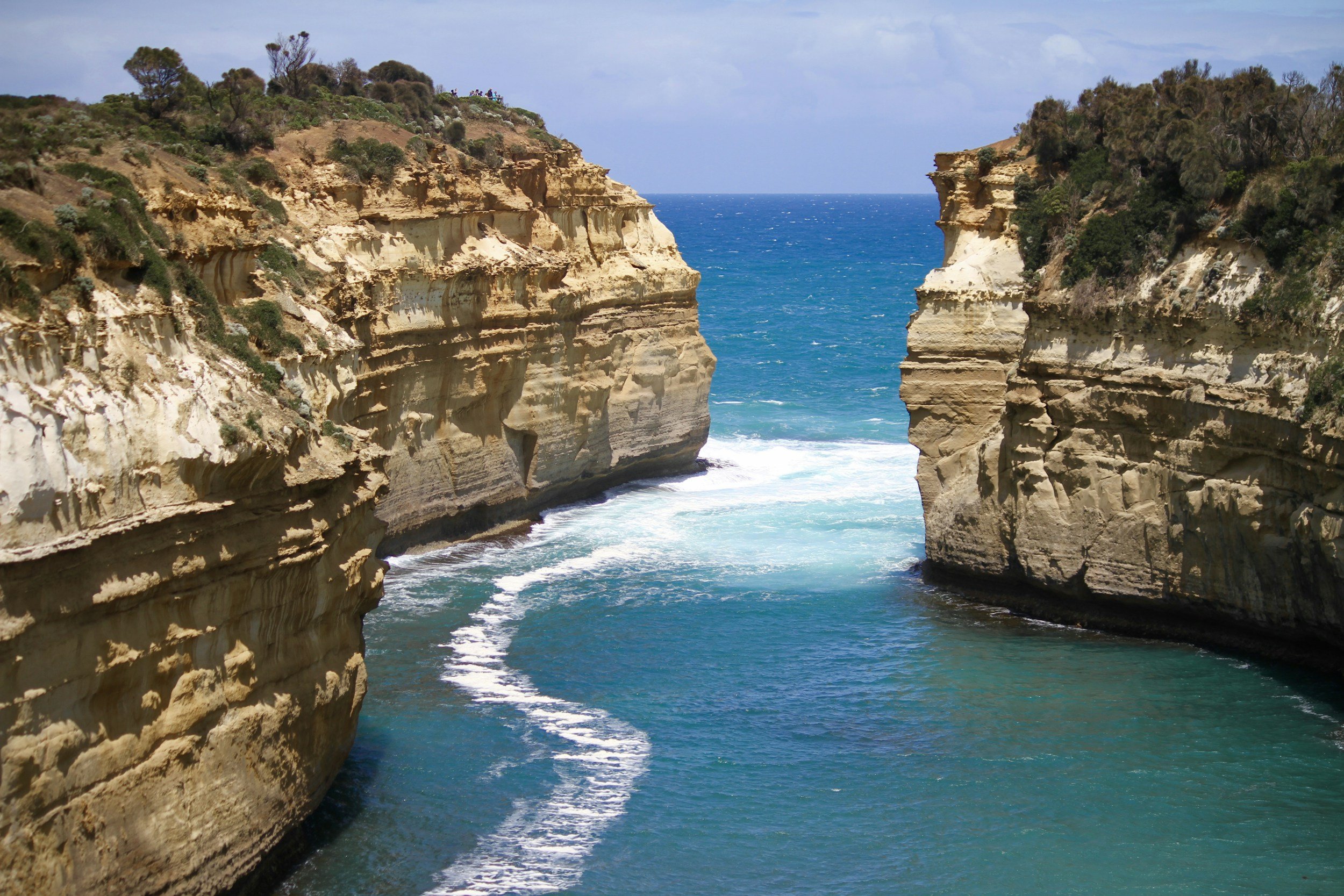 Cliffs overlooking the ocean with people standing at the top, beneath a partly cloudy sky.