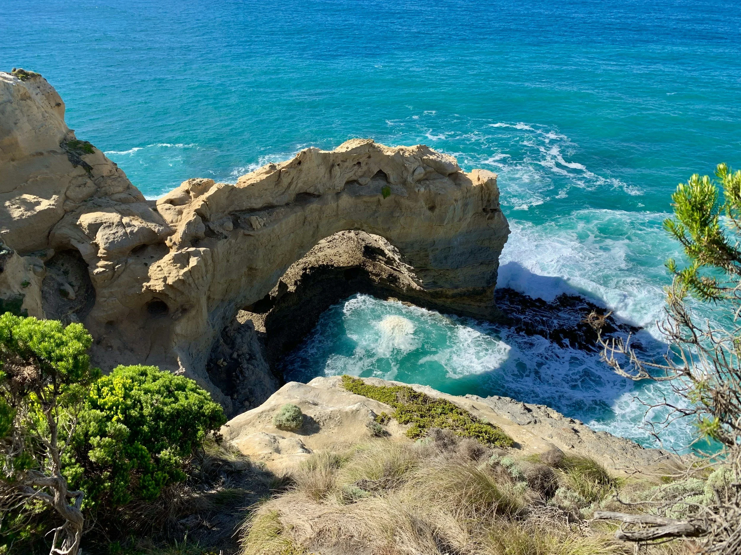 Cliffside view with turquoise ocean water, rocky formations, green shrubbery, and blue sky in the background.