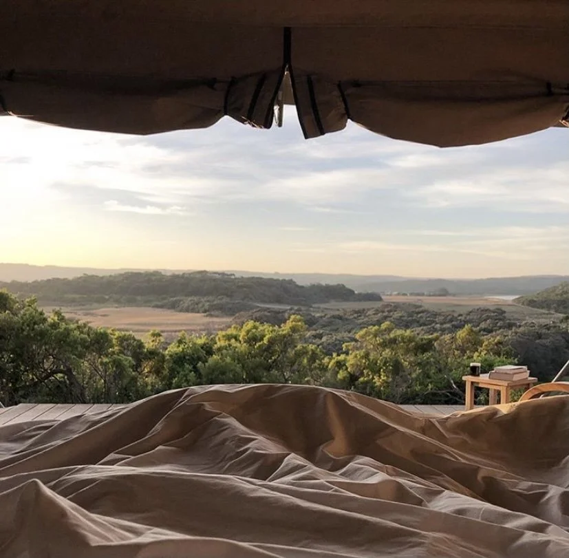 View of a scenic landscape with rolling hills, trees, and a river during sunset, seen from a bed under a canopy with a table and books nearby.
