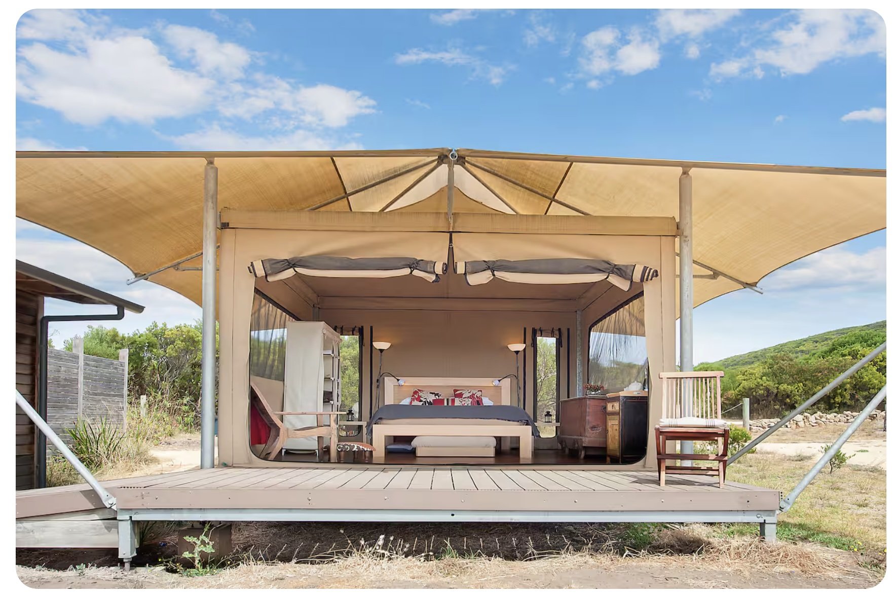 A cozy outdoor bedroom with a bed, side tables, and a beige canopy overhead, set on a wooden deck with a scenic backdrop of green hills and blue sky.