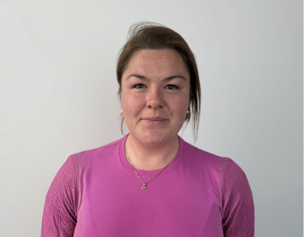A woman with short brown hair wearing a pink top, standing against a plain white background.