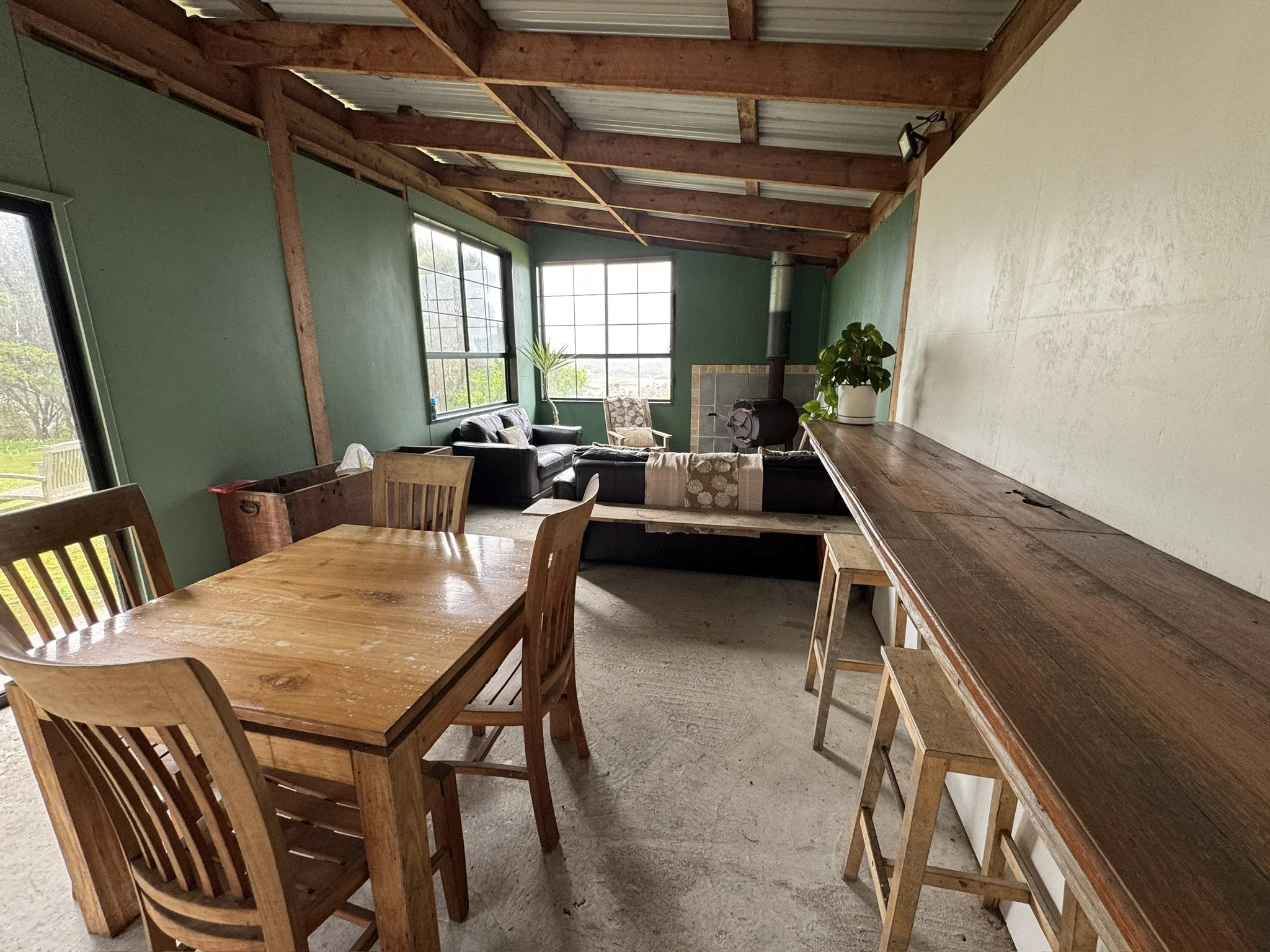 The image shows the interior of a rustic living space with exposed wooden beams on the ceiling and green painted walls. There are large industrial-style windows letting in natural light. The room contains a black leather sofa, a wooden lamp, a potted