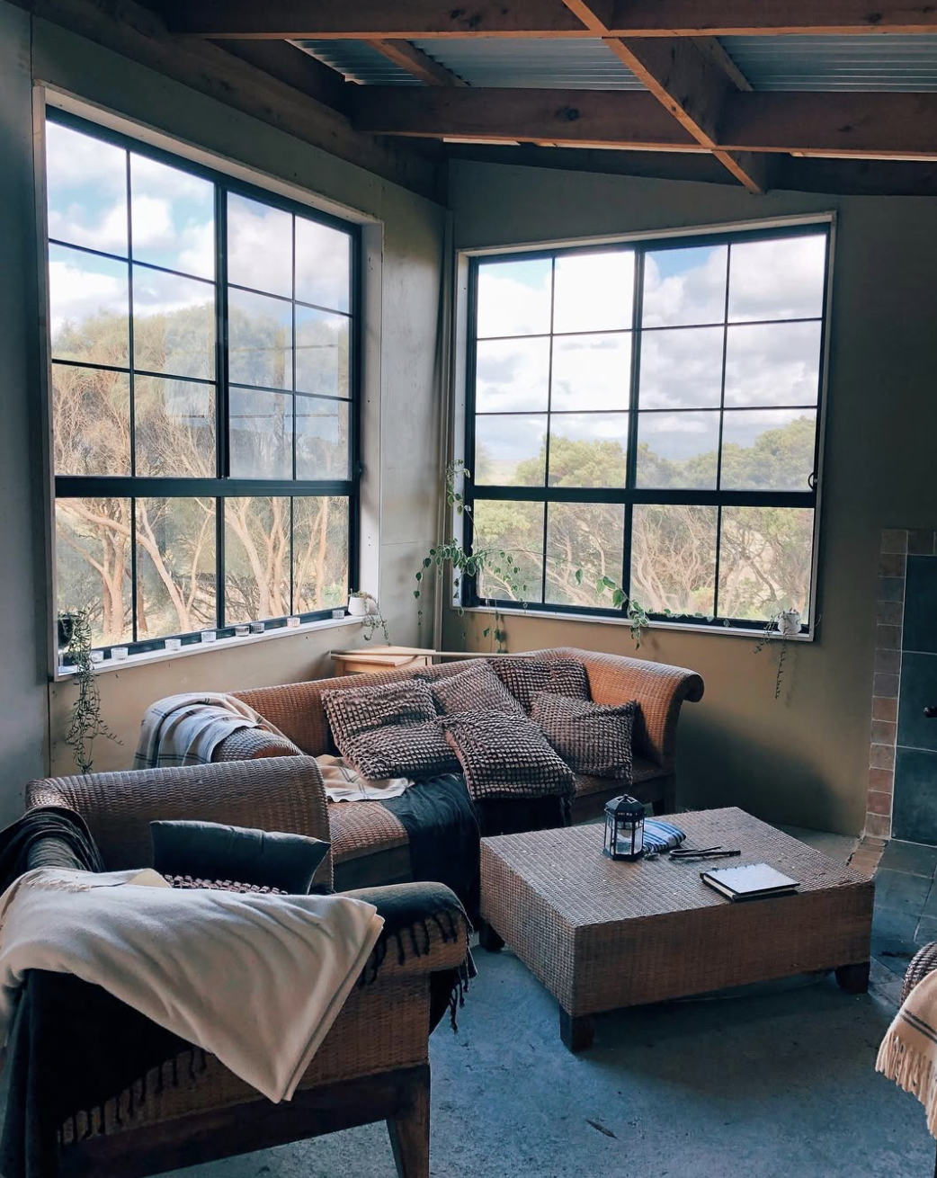Living room with large windows showing trees outside, a wicker sofa with patterned pillows, a wicker armchair with a blanket, a rattan coffee table with a lantern, notebook, and sunglasses, and potted plants.
