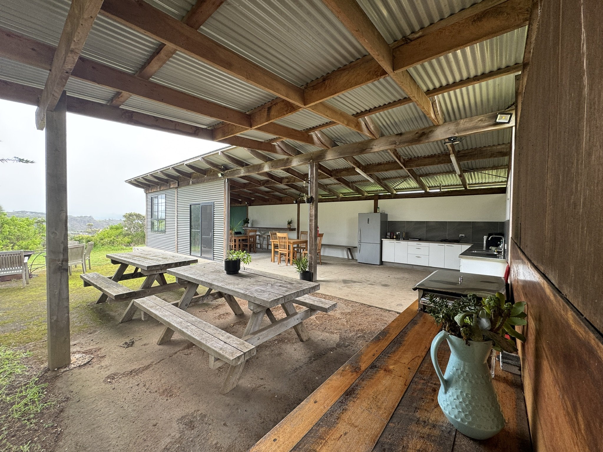 An outdoor covered patio area with wooden beams and a corrugated metal roof, featuring a rustic wooden picnic table and benches, potted plants, and a kitchen area with white cabinets and a countertop.