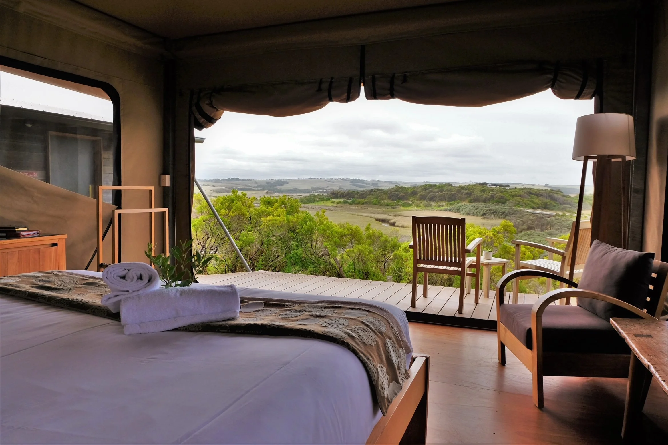 A cozy bedroom overlooking a scenic landscape of green hills and trees through an open sliding door. The bed has neatly folded towels and a blanket, with some furniture including chairs and a floor lamp nearby.