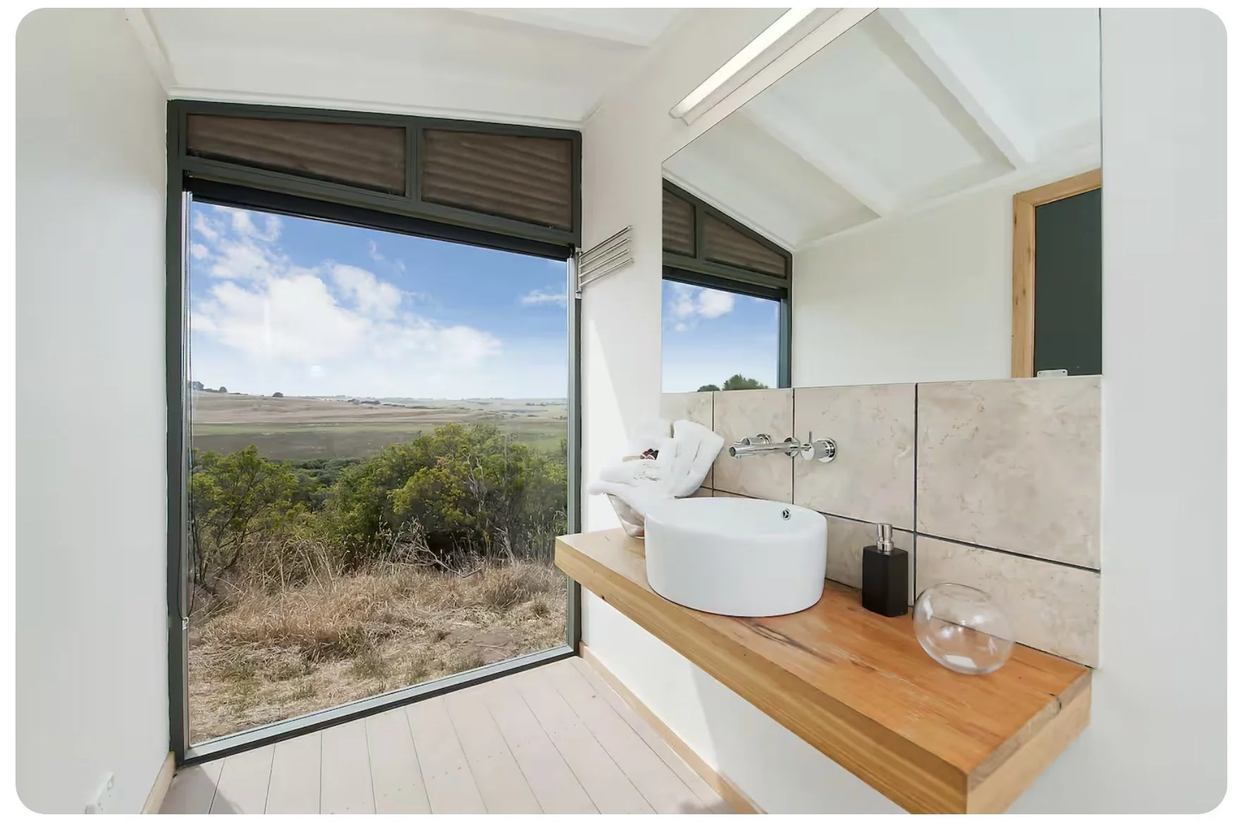 Bathroom with a large window showing a landscape of open fields and trees, a white vessel sink on a wooden countertop, and a partial view of a mirror and soap dispenser.