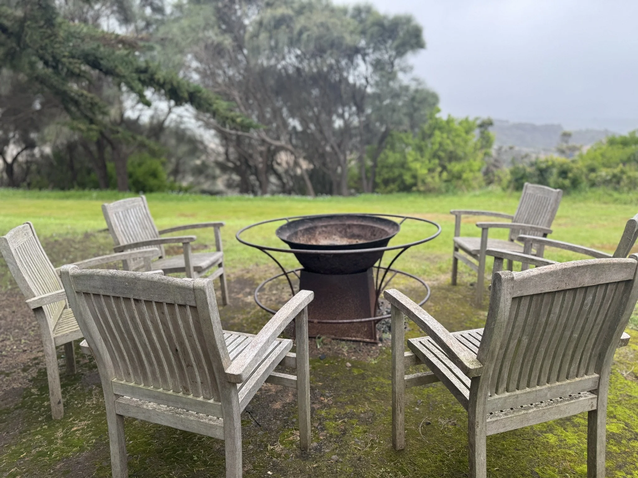 A round metal fire pit surrounded by eight weathered wooden outdoor chairs on a grassy area with trees in the background, under an overcast sky.