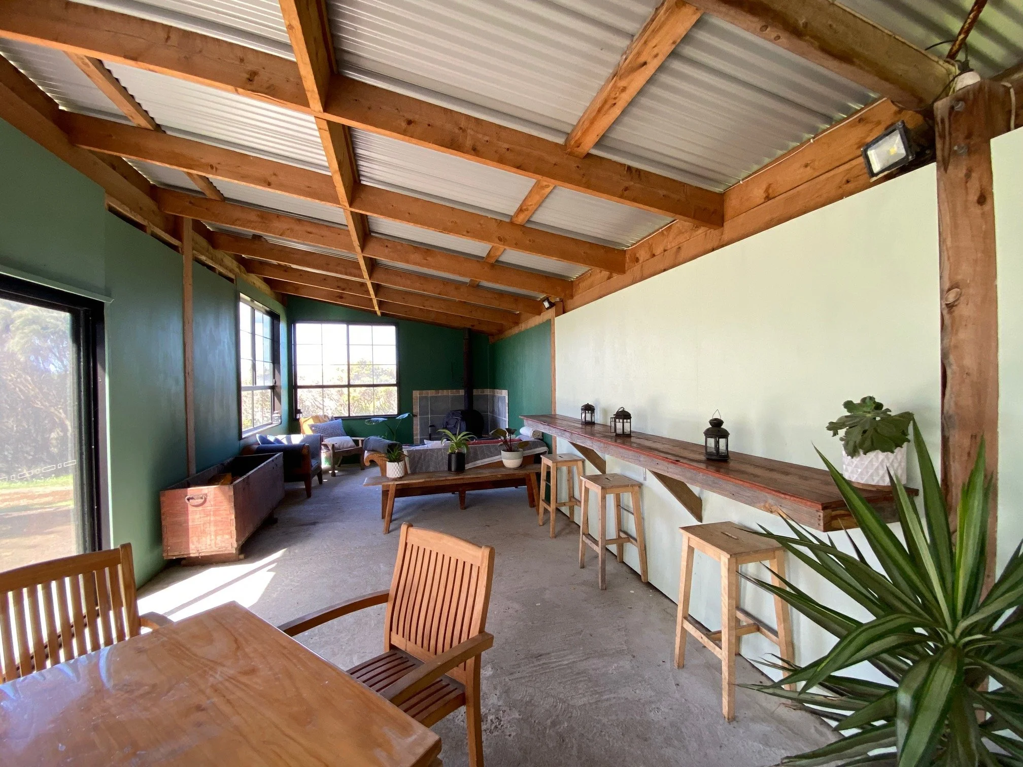 Interior of a room with a wooden ceiling, green walls, large windows, a wooden dining table with chairs, a lounge area with couches, a fireplace, and decorative plants.