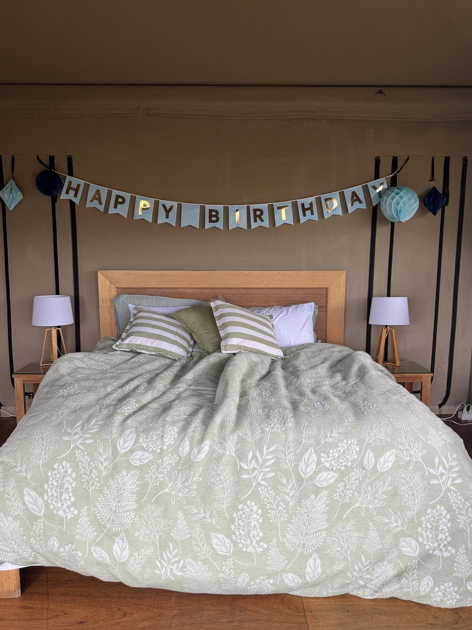 A bedroom with a bed covered in a light green and white floral-patterned bedspread, two striped pillows, two lamps on wooden nightstands, and a 'Happy Birthday' banner hanging above the headboard, decorated with paper pom-poms and hanging decorations