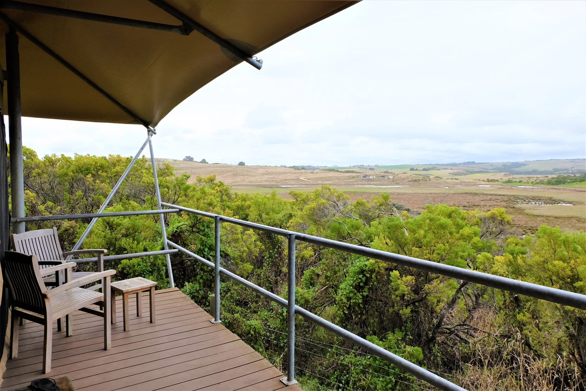 A balcony with wooden chairs and a railing overlooking green trees and open fields with hills in the distance under a cloudy sky.