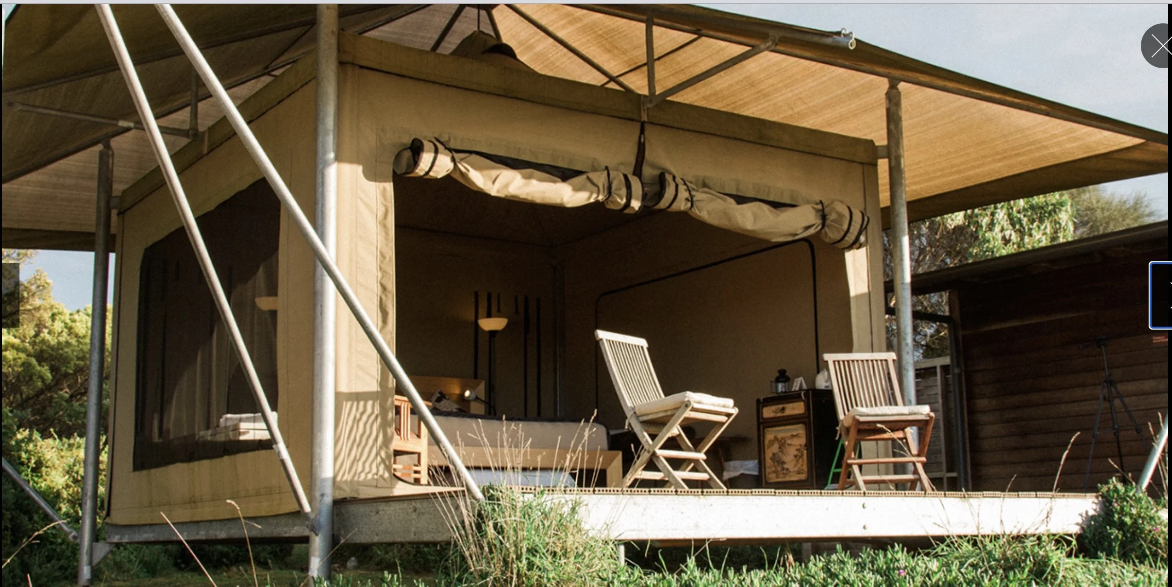 A small house or shed under construction with scaffolding around it. The house has a screened porch with patio furniture and an open doorway. The roof is in the process of being built, with unfinished exterior. There are trees and grass in the surrou