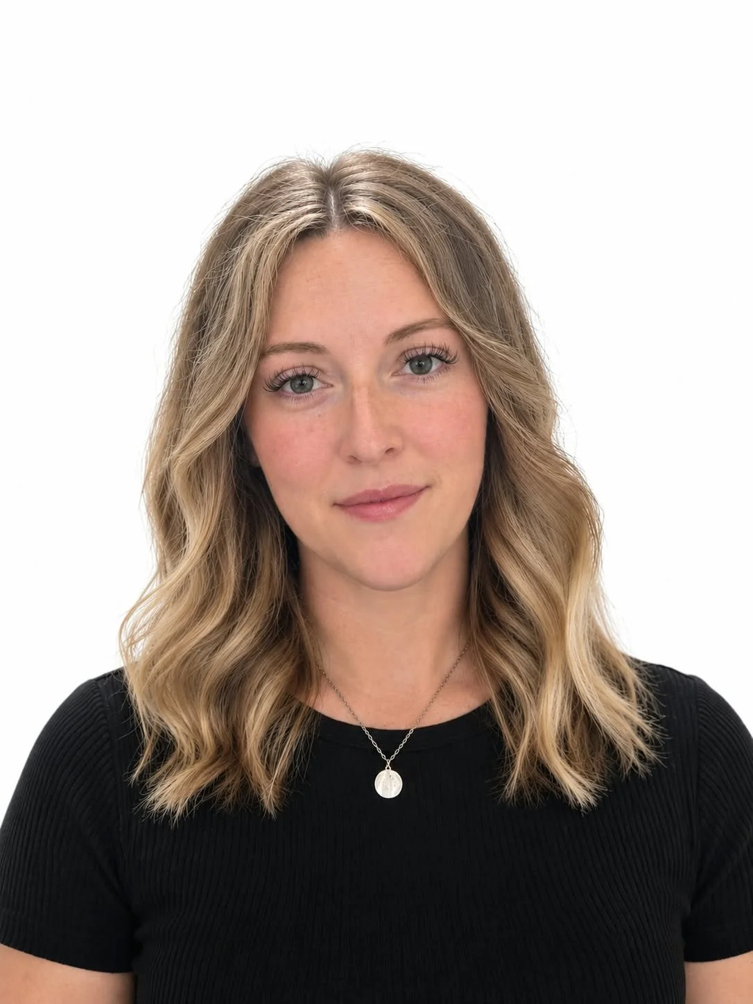 A woman with shoulder-length wavy blonde hair, wearing a black top, a silver necklace with a round pendant, and light makeup, against a white background.