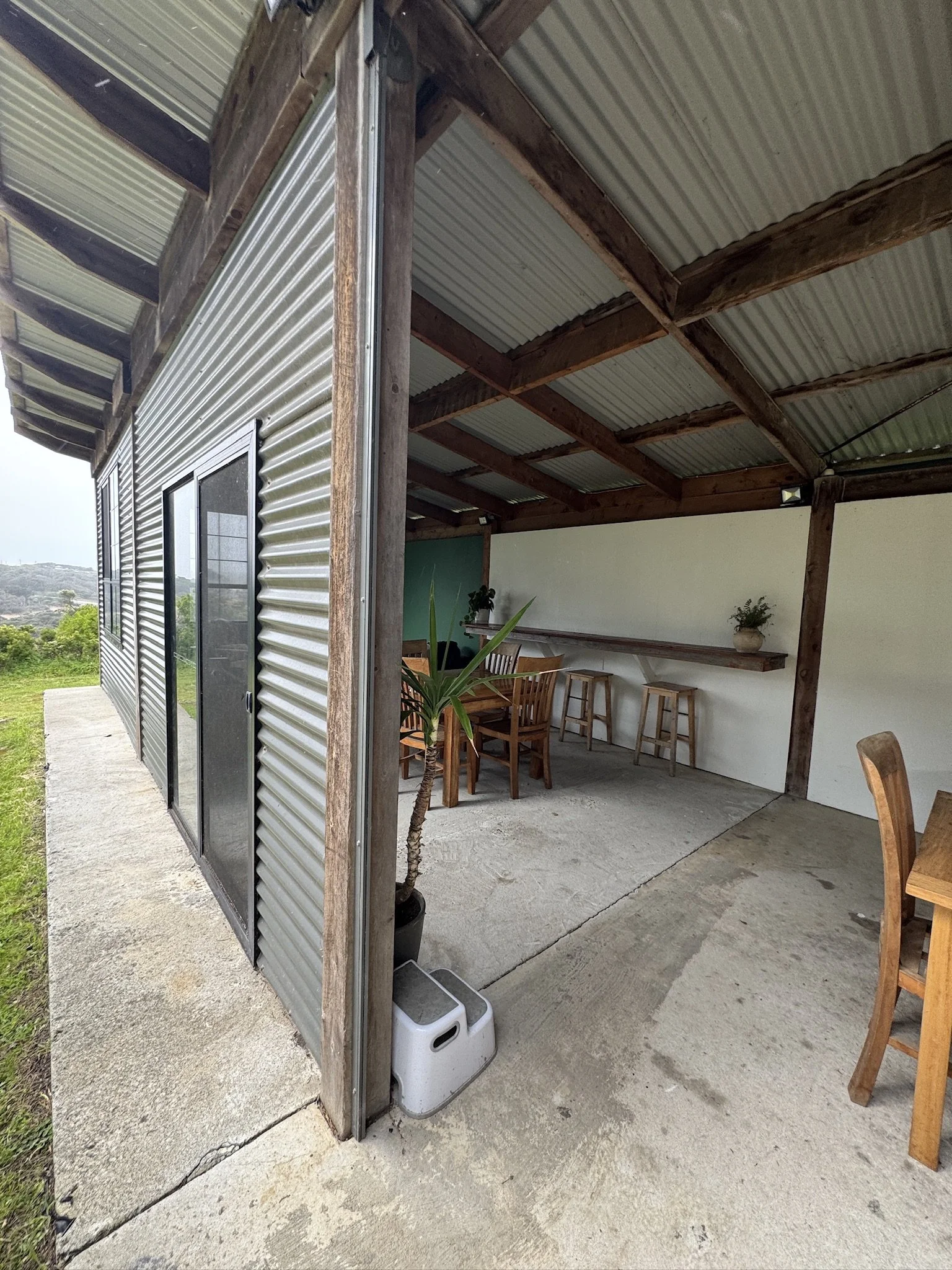 An outdoor patio area with a corrugated metal roof and wooden beams, partially enclosed with a wall and large window. Potted plants and wooden chairs are visible, along with a small air conditioning unit on the concrete ground outside.