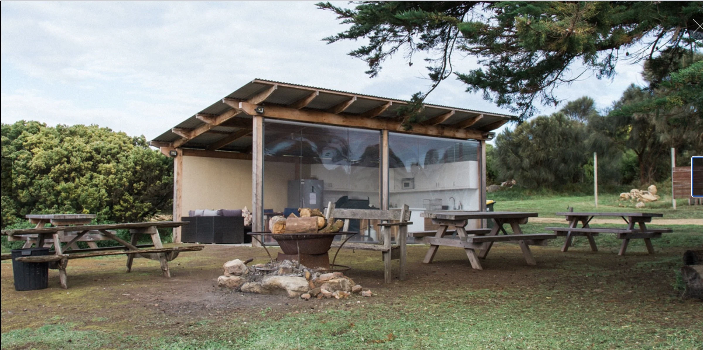 An outdoor patio area featuring several wooden picnic tables, a fire pit with rocks, and a small building with a glass front and a corrugated metal roof, surrounded by greenery and trees.