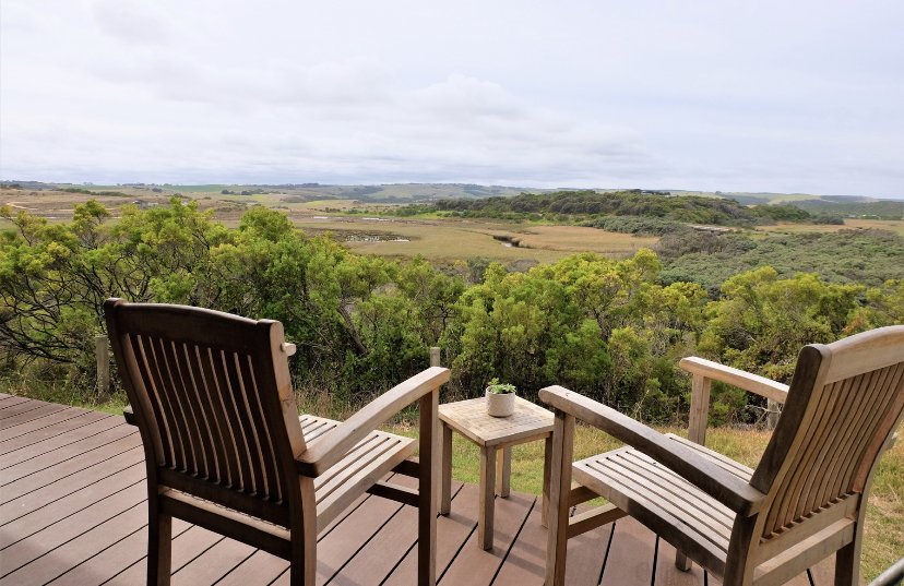 Two wooden chairs and a small table with a plant on it on a deck overlooking a lush green landscape with trees and open fields under a cloudy sky.