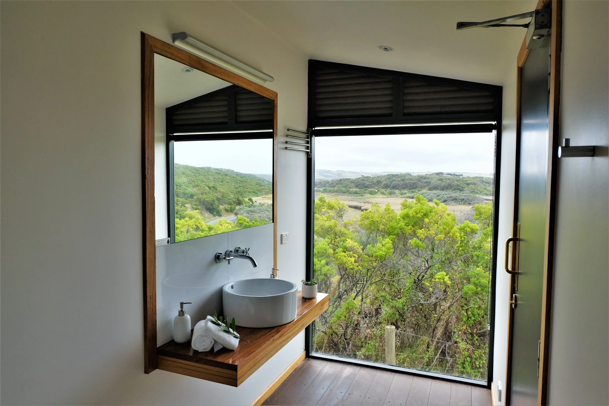 Small bathroom with a window showing a green landscape outside. The bathroom has a white round vessel sink on a wooden shelf with rolled towels, a soap dispenser, and a mirror above the sink. There is a bottle of soap next to the sink. The window has black blinds and a view of trees and rolling hills.