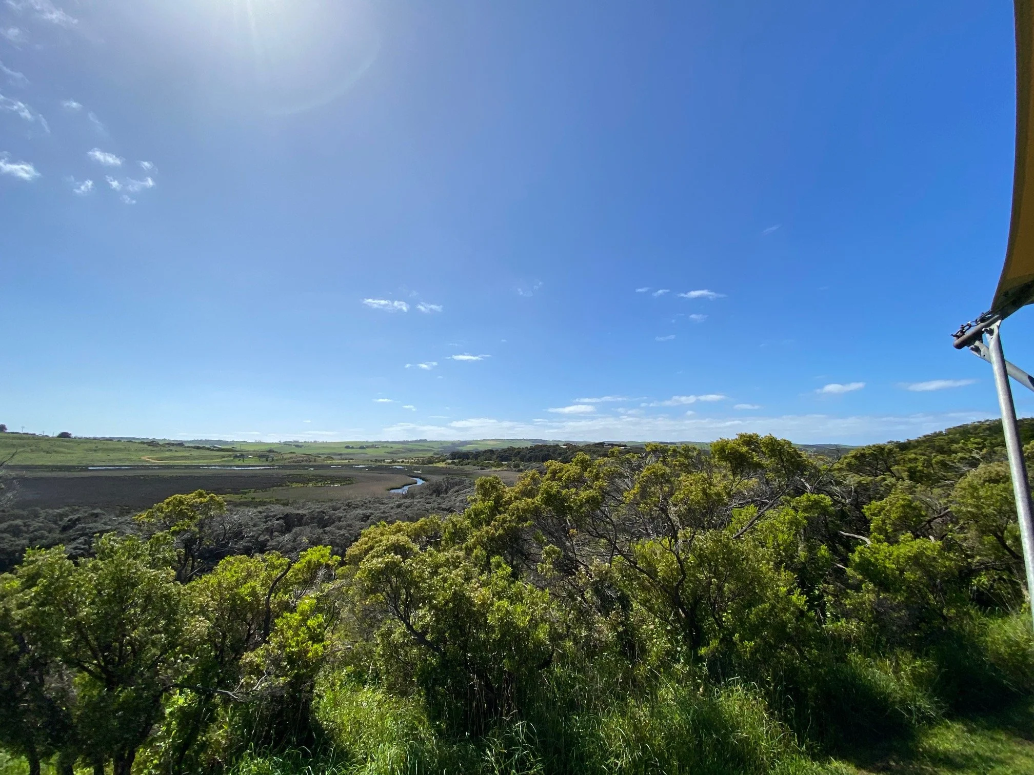 A bright blue sky with a few small white clouds and a partial view of a yellow structure on the right side, overlooking a lush green landscape with trees and water.