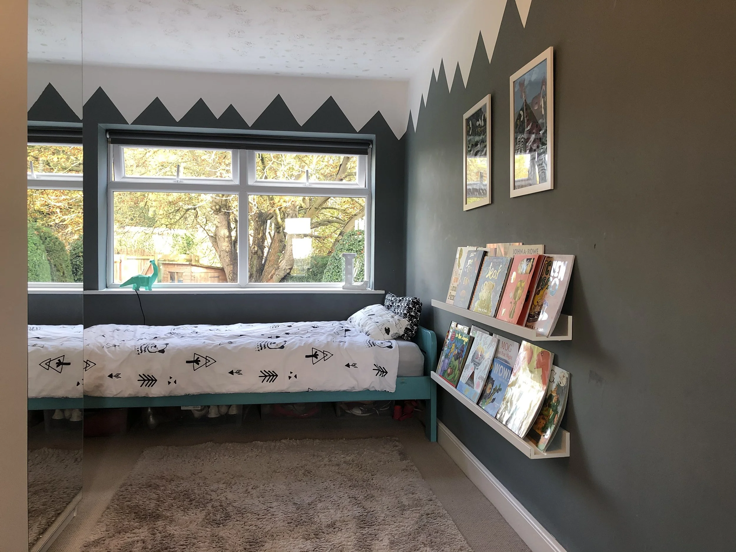 A bedroom with a large window showing trees outside. The bed has a black and white patterned bedding, and next to it is a white bookshelf filled with children's books, with some books leaning against the wall.
