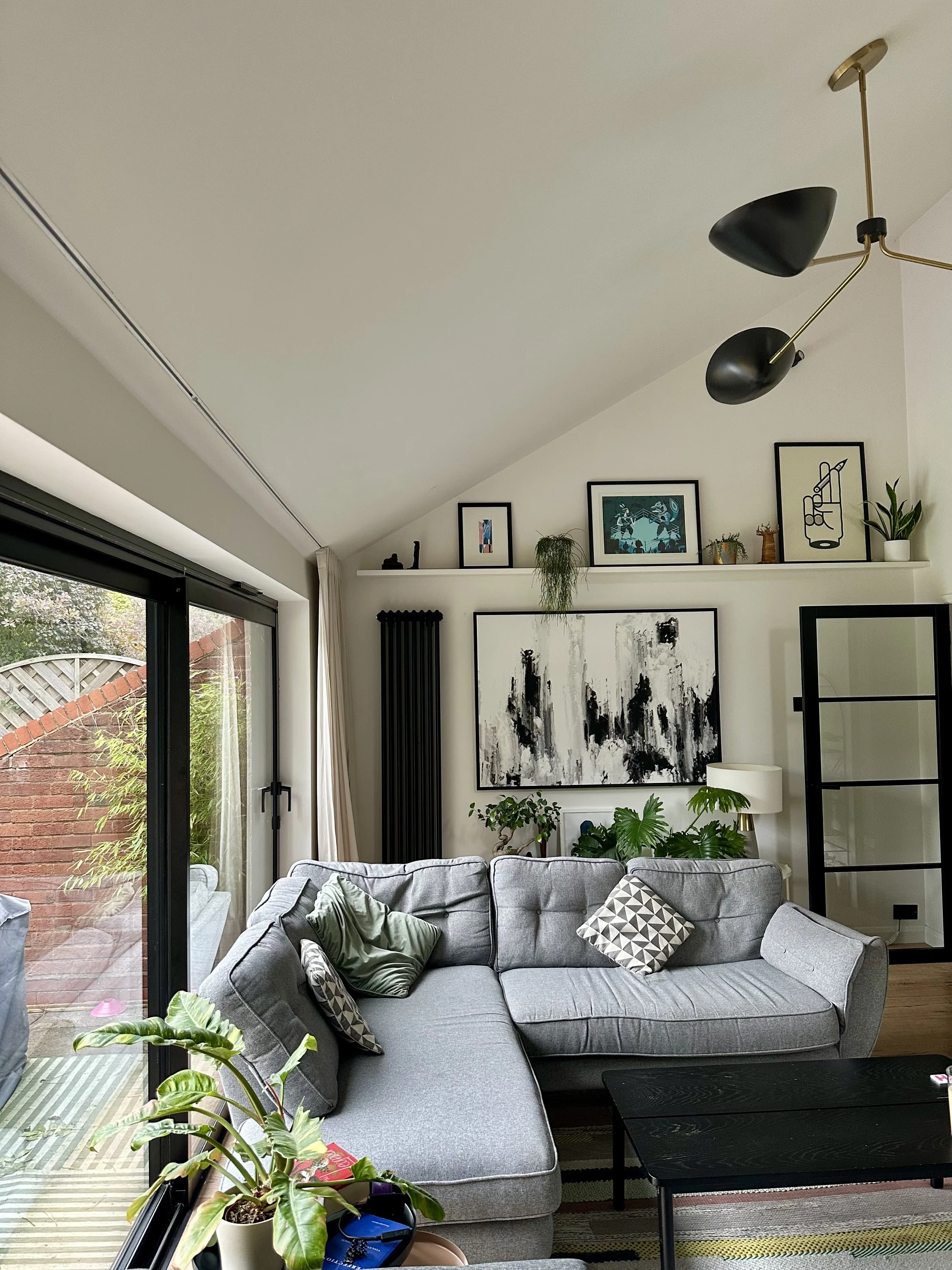 Living room with gray sectional sofa, black coffee table, artwork on the wall, large window with sliding door leading to a patio, plants near the window, and contemporary ceiling light fixture.