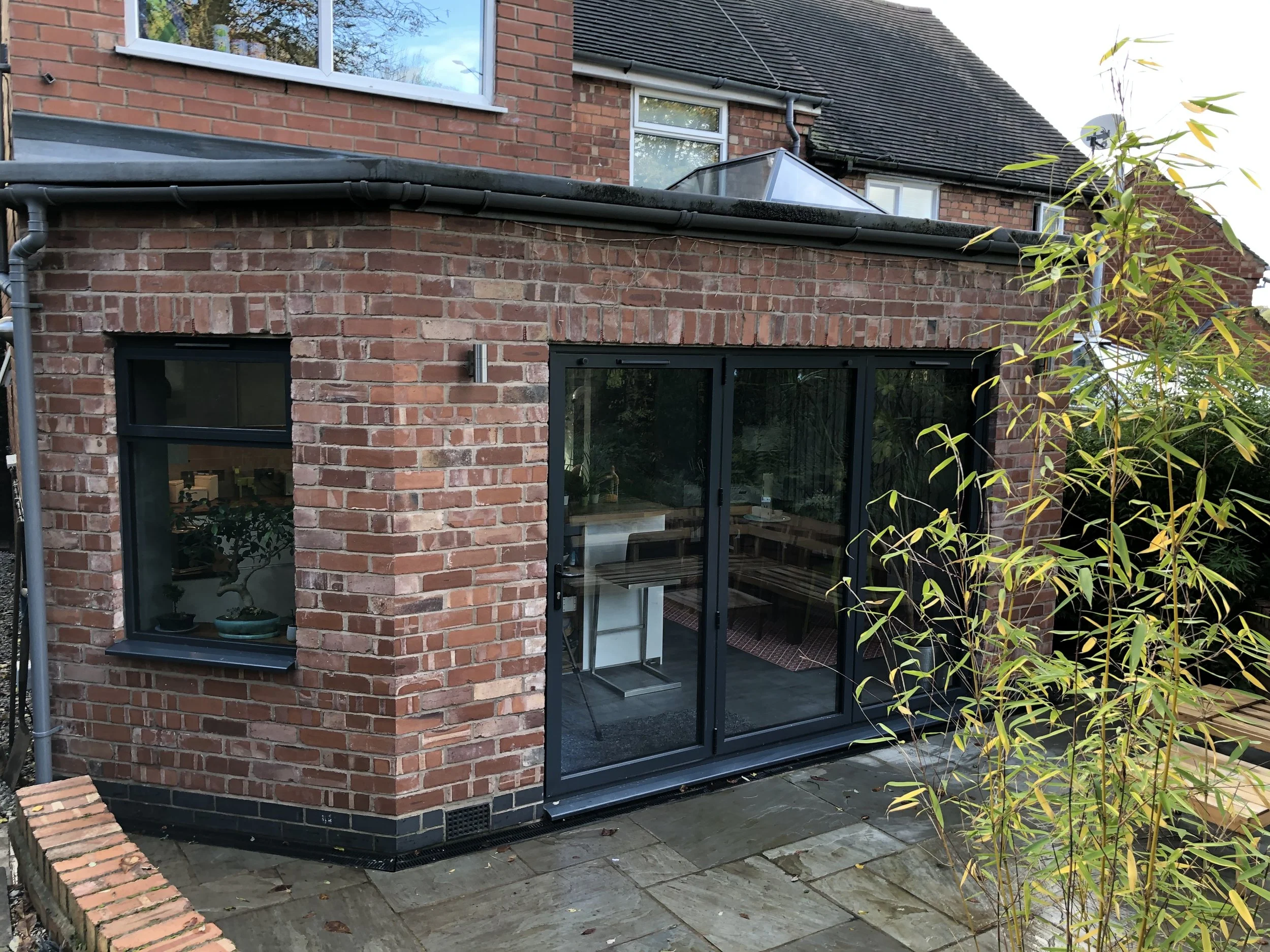 The photo shows a part of a brick house with large glass doors and windows, and a small brick wall in front. There is a corner of a patio with paving stones and some greenery, including a small shrub with yellow-green leaves on the right side.