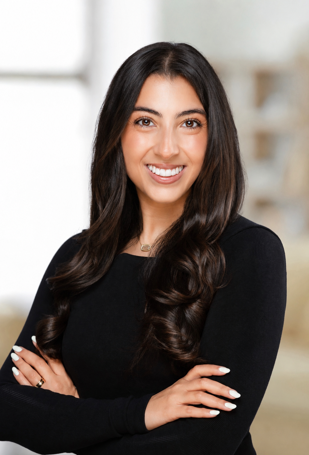 A young woman with long dark hair, wearing a black top, smiling with crossed arms in a bright modern interior.