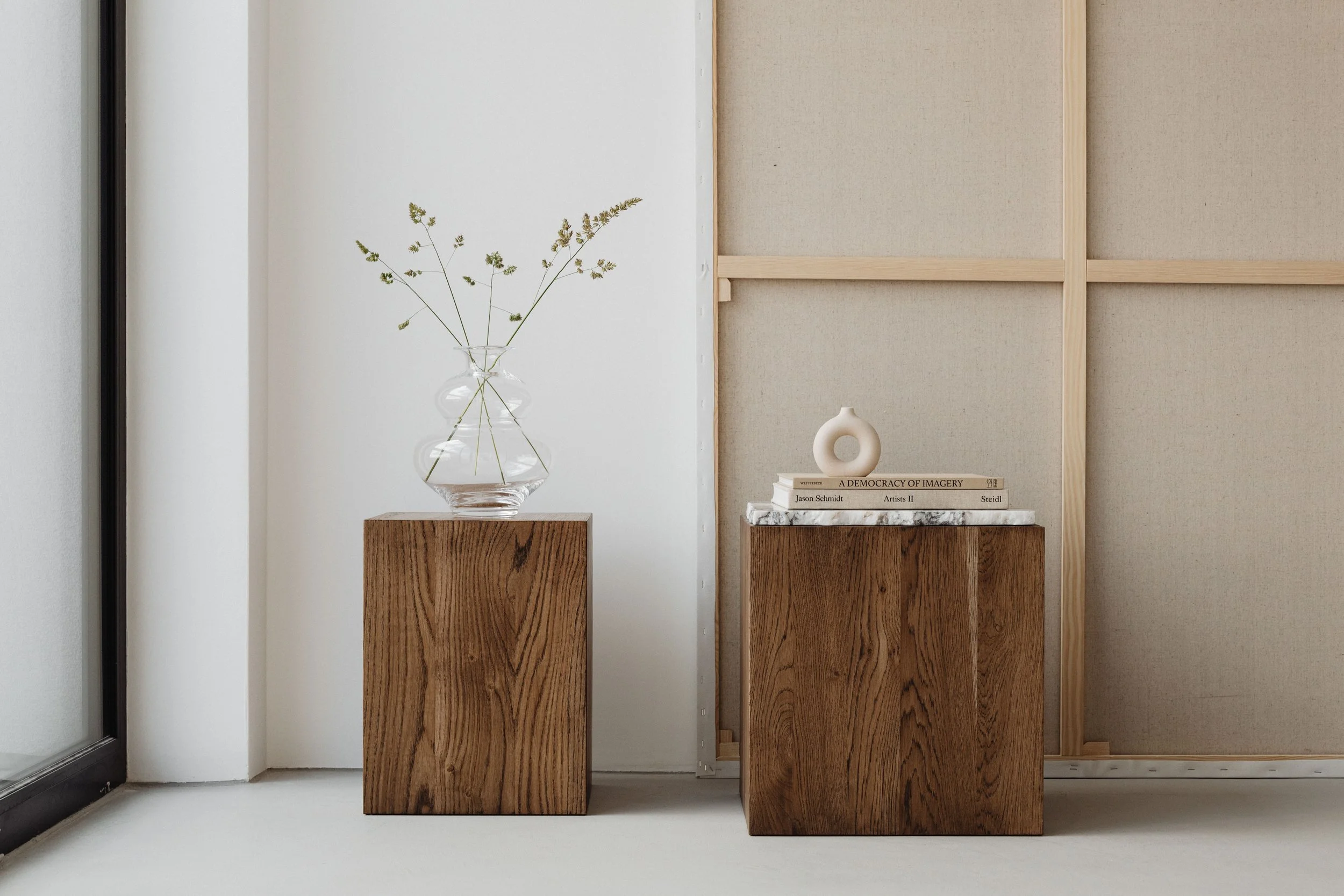 Minimalist interior with three wooden pedestals, a glass vase with dried greenery, and a stack of books including "A Democracy of Imagery" by Jason Schmidt on a marble surface. Behind, a textured beige wall section and part of a grey wall are visible.
