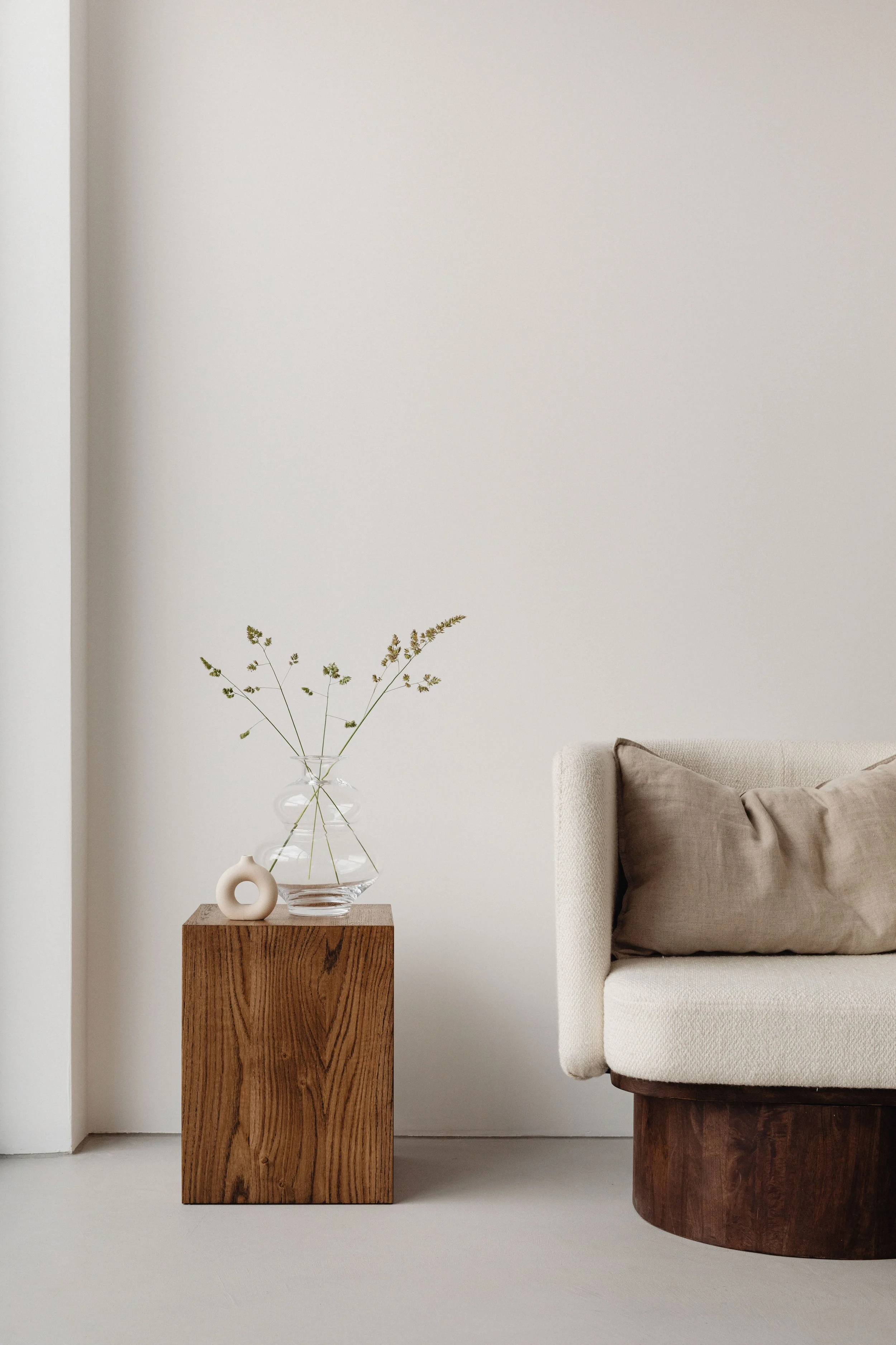 A beige sofa with a brown cushion next to a wooden block used as a side table, which has a clear glass vase with green plants and a small white decorative object on it, against a plain light wall.