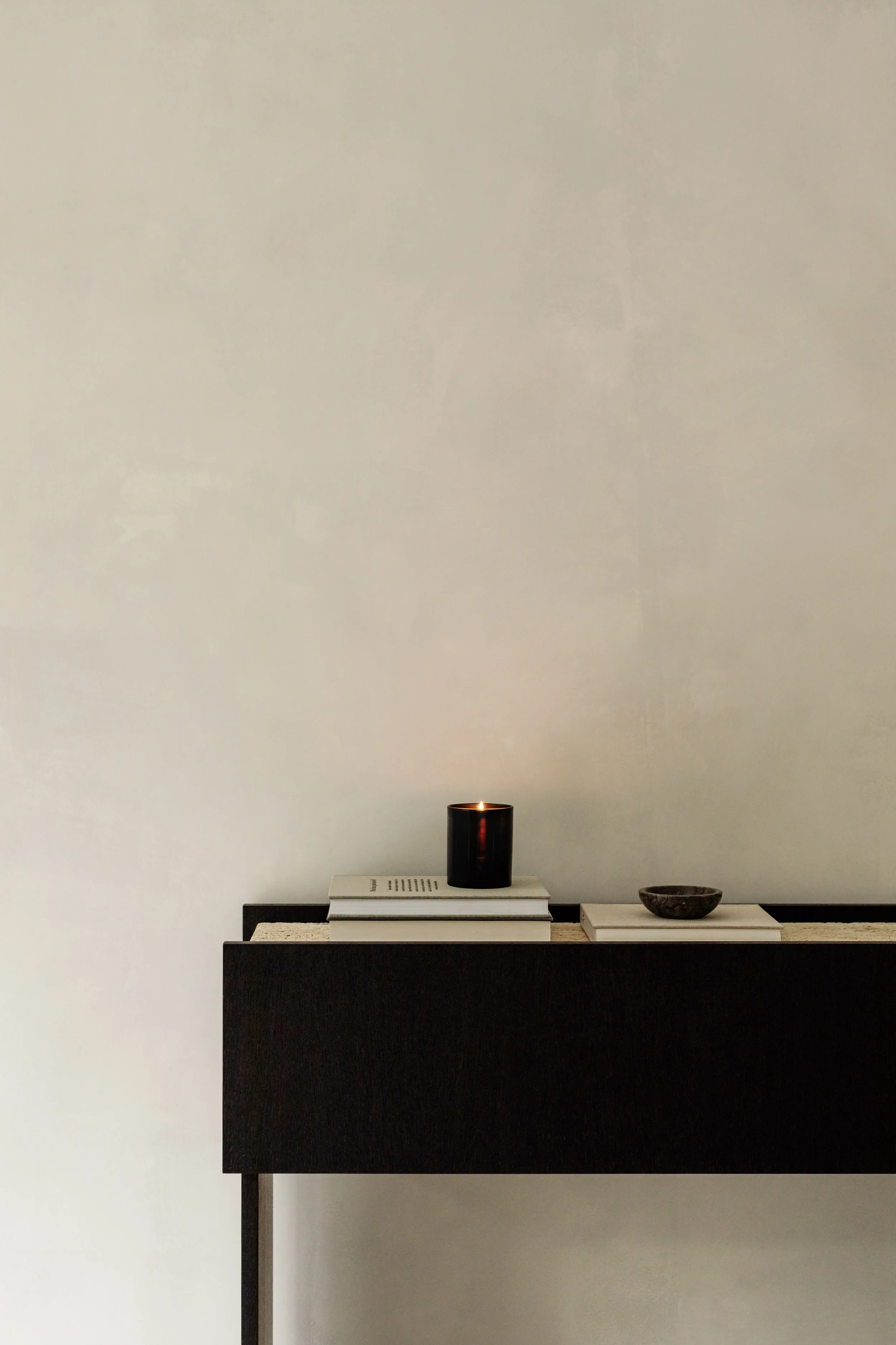 A black candle is lit, placed on top of a stack of white books on a modern black and beige sideboard in a minimalist room with plain white walls.