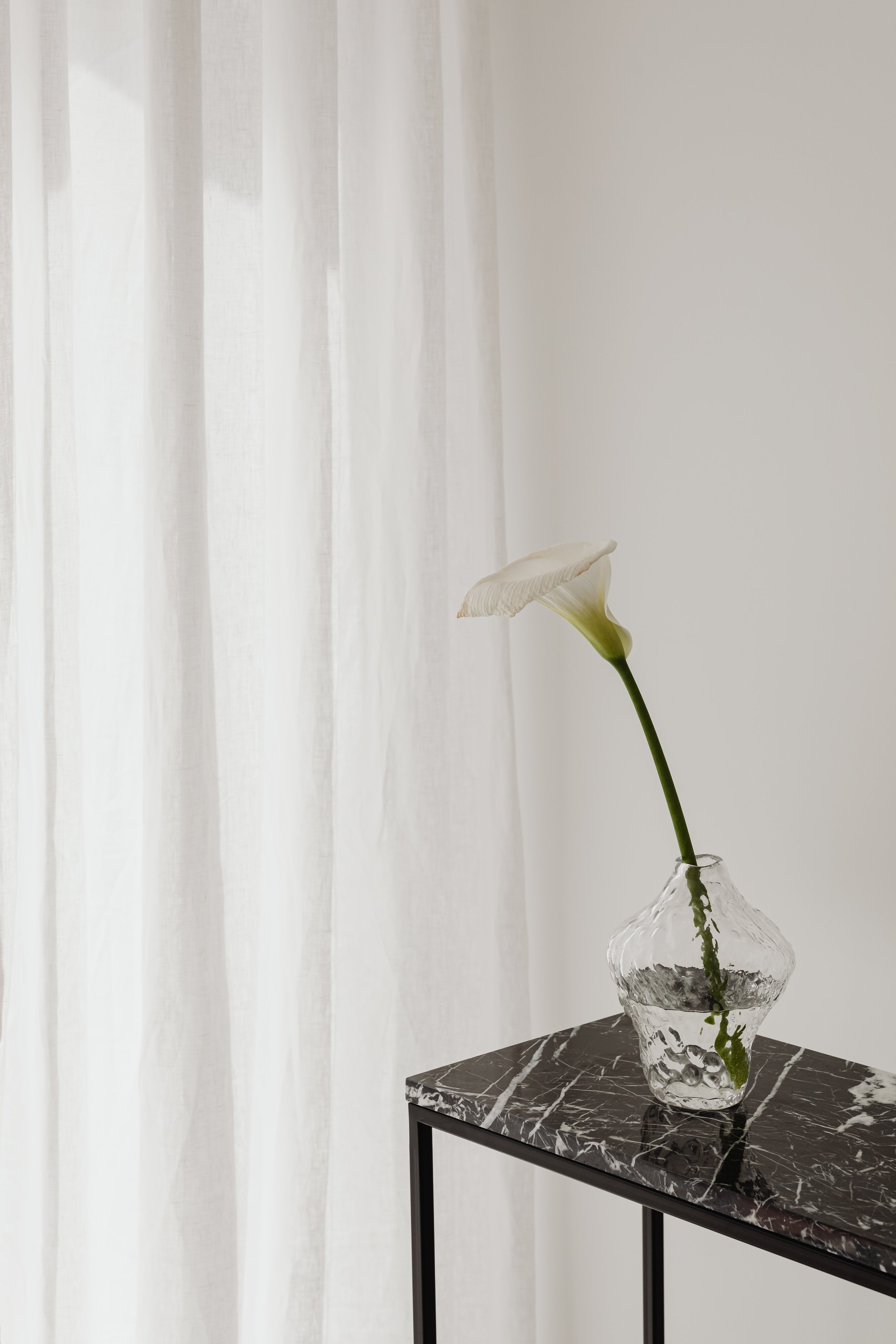 A single white flower with a curved petal in a clear, textured glass vase on a black marble-topped side table. White curtains are in the background.