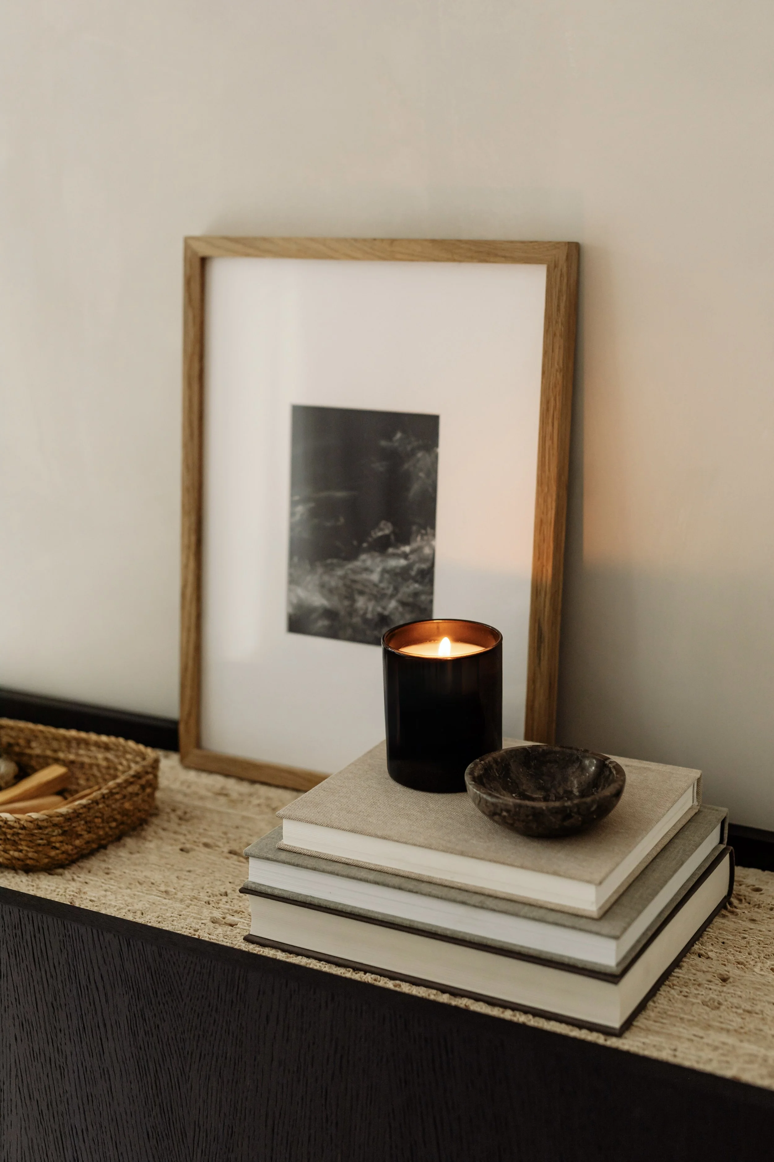Decorative table setup with stacked white books, a black candle, a small dark bowl, and a picture frame with black and white artwork behind.