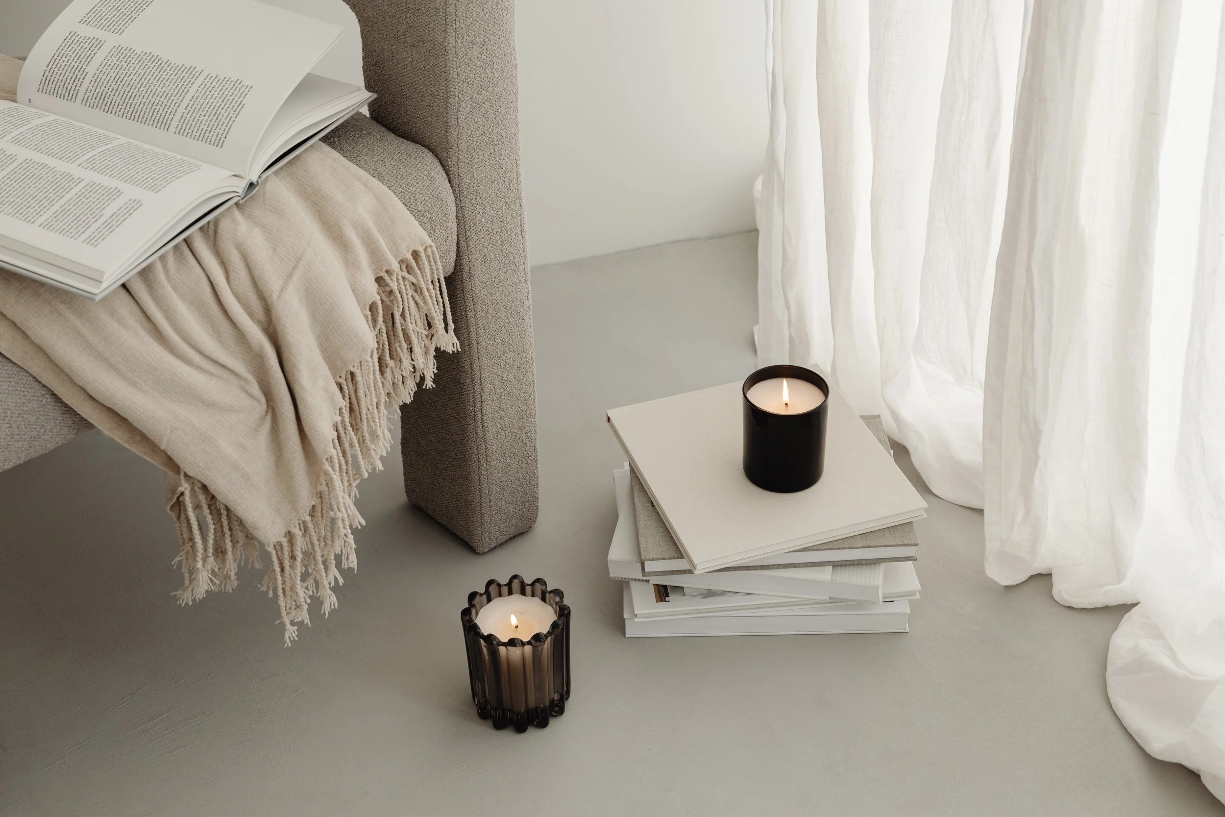A cozy corner with a beige upholstered bed, a soft beige throw blanket, an open book on the bed, a white coffee table stacked with books, two black candles with flames, and sheer white curtains letting in natural light.