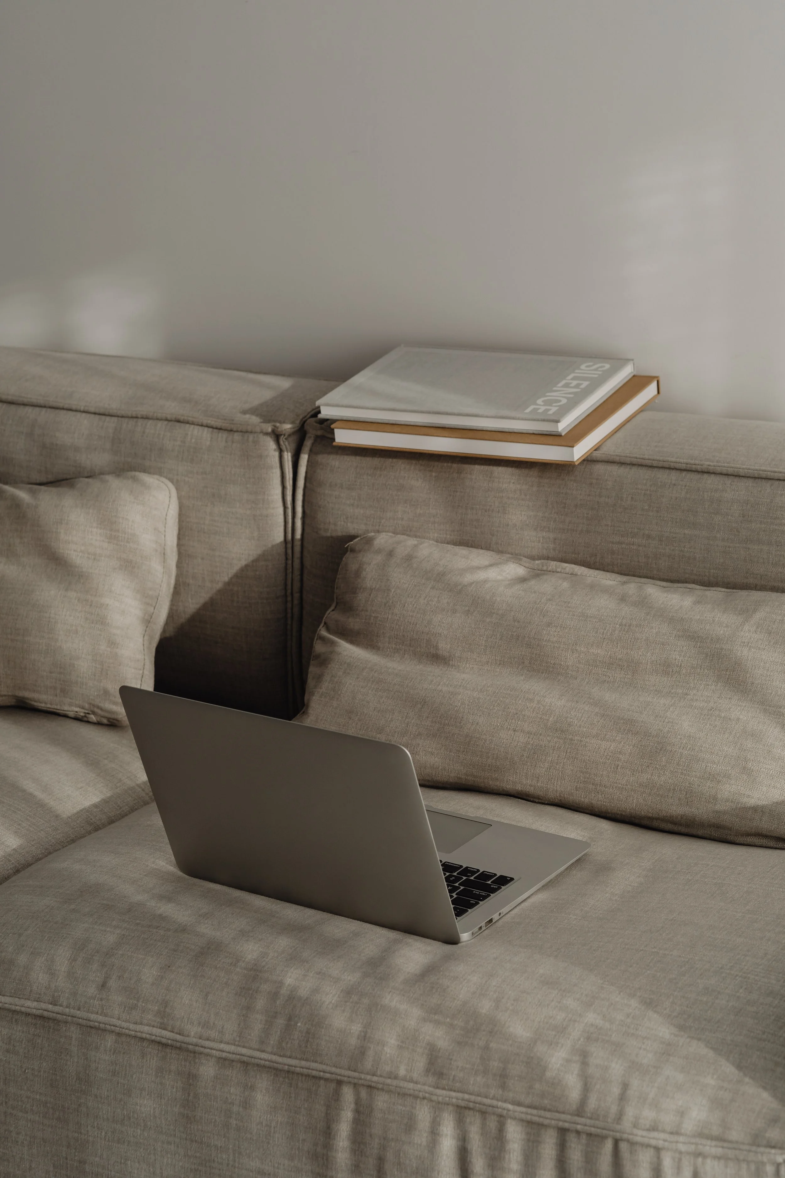 L-shaped beige sofa with cushions, a gray closed laptop, and two stacked books on the armrest, with a light-colored wall in the background.