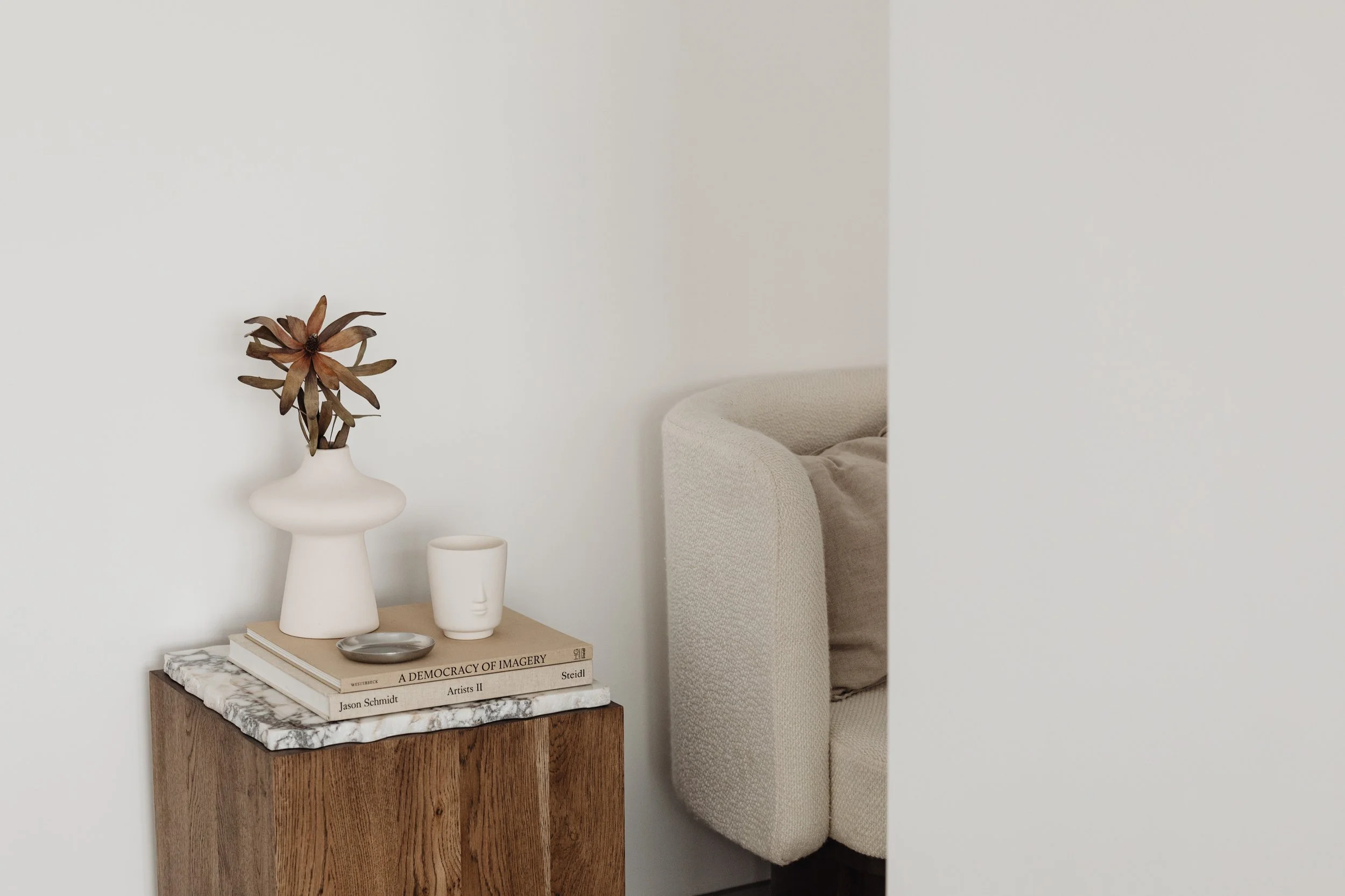 A small wooden side table with a marble top, decorated with a stack of books, a small silver dish, and white sculptural vases holding dried brown flowers, set against a plain white wall and partially visible cream-colored armchair.
