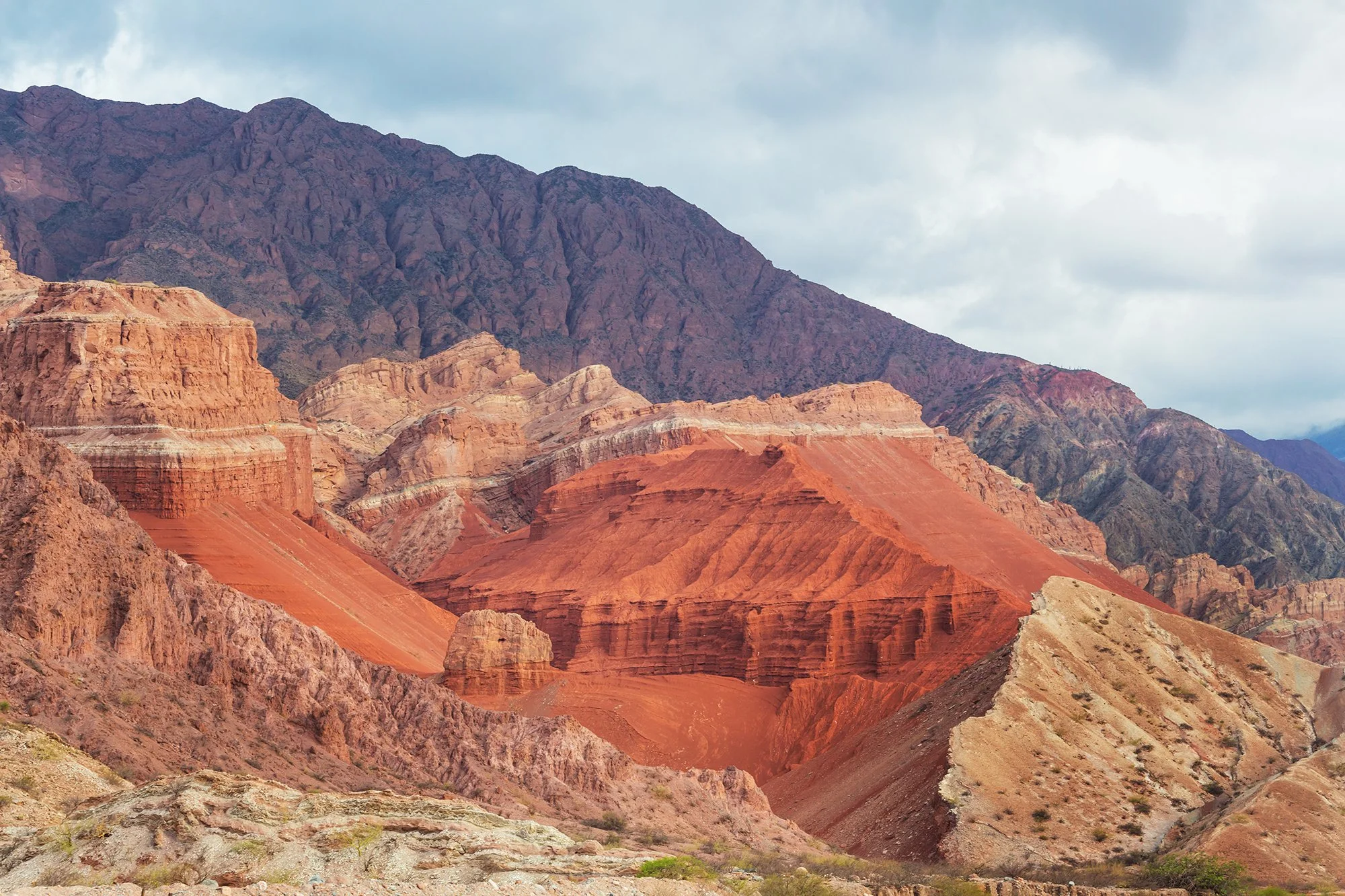 Colorful layered rock formations and mountains under a cloudy sky.