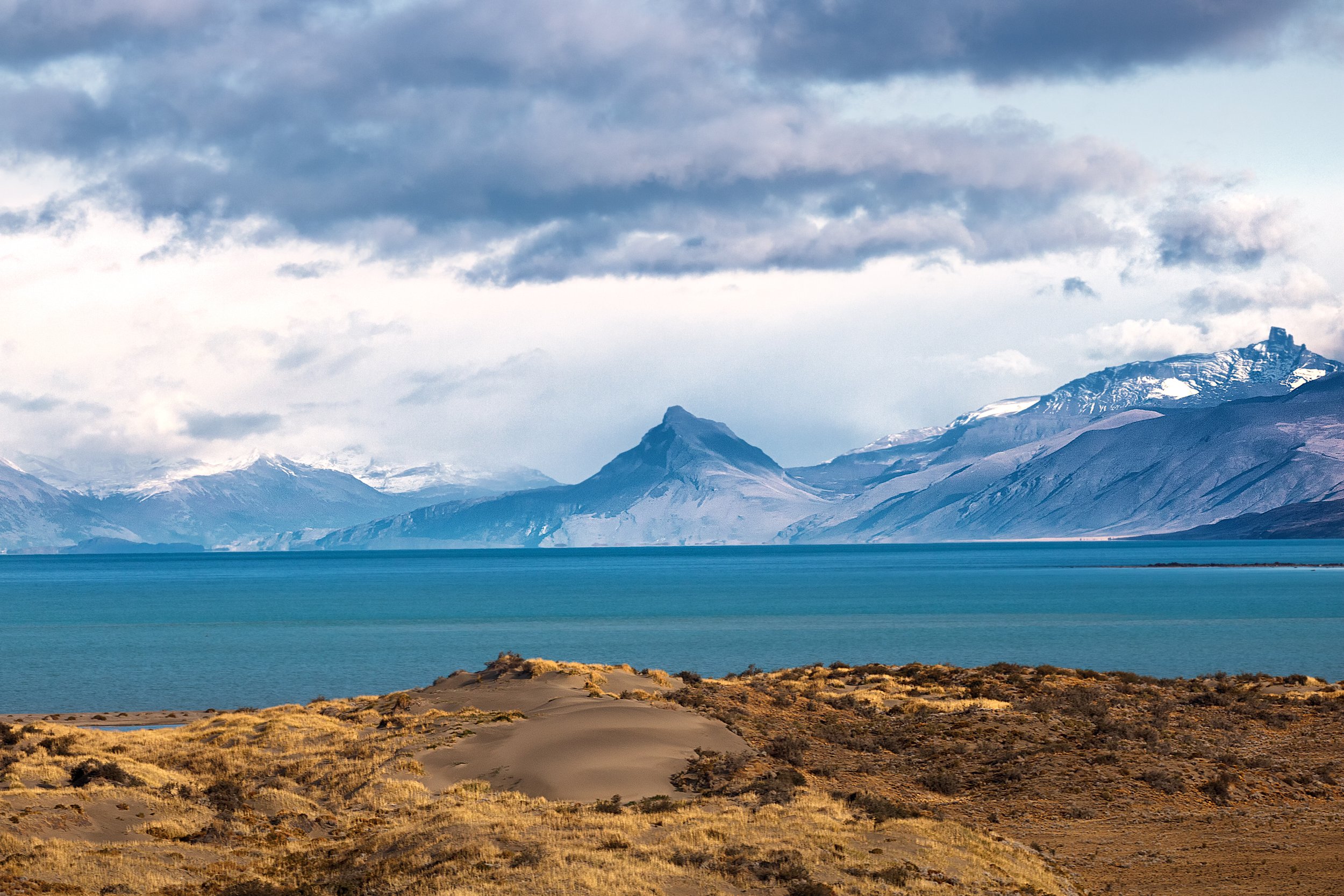 Scenic view of a large body of water with mountains in the background, partially snow-capped, under a cloudy sky. Foreground shows sandy and grassy terrain.