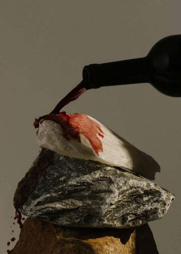 A wine bottle pouring red wine onto a rock and a white stone, with a third larger rock underneath.