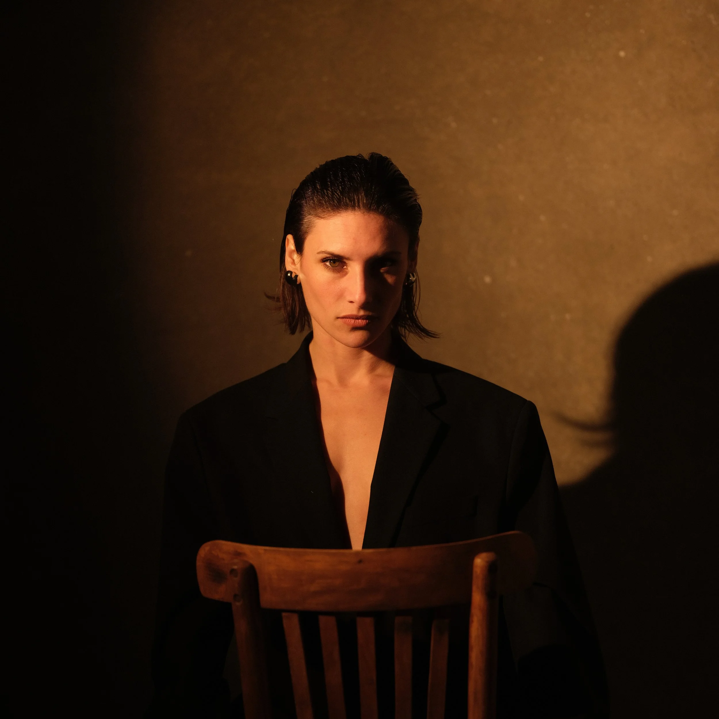 Mujer con cabello oscuro y peinado hacia atrás, usando chaqueta negra, en un fondo oscuro, sentada en una silla de madera.
